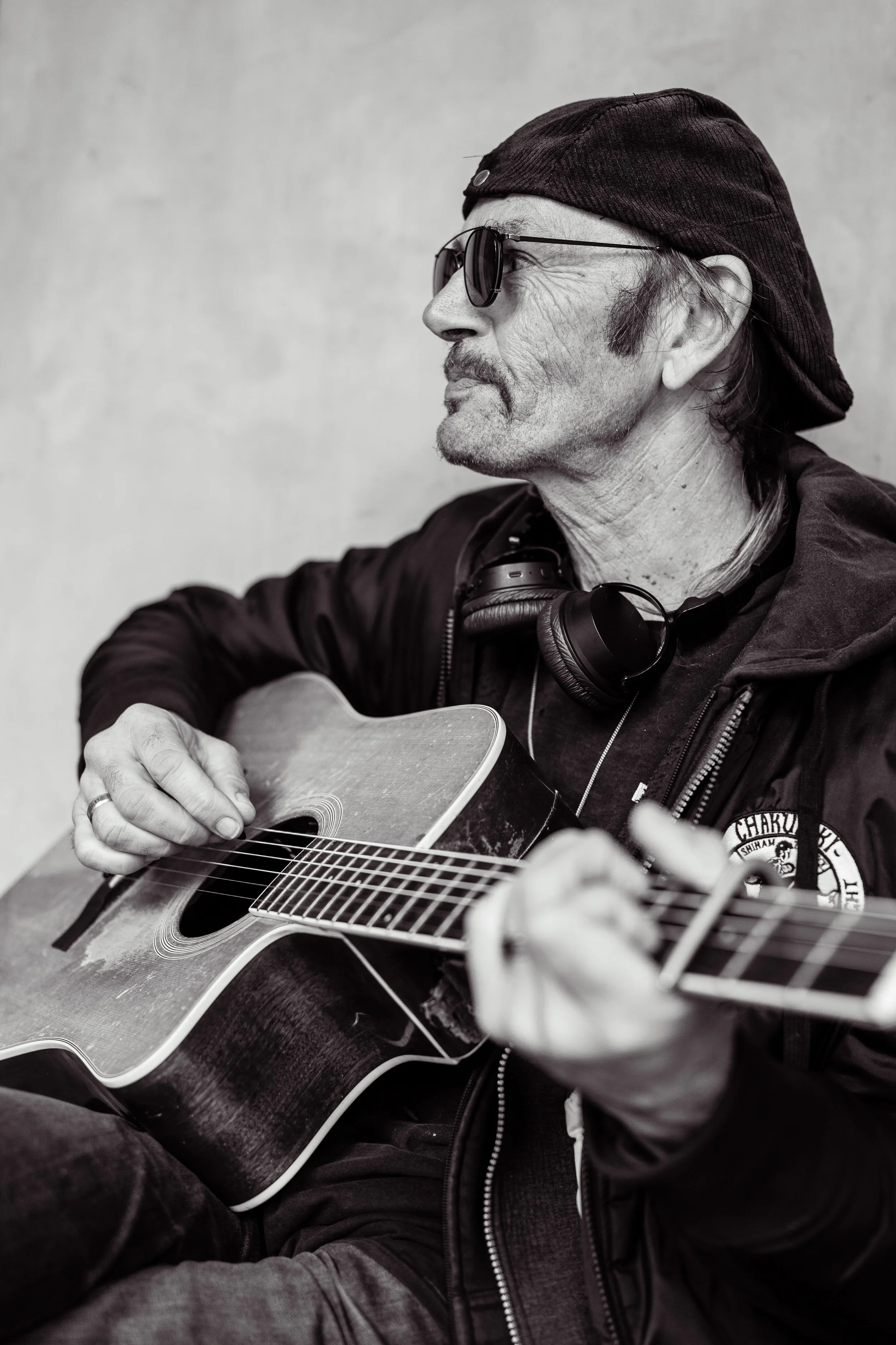 Een man met een pet, zonnebril en headphones speelt gitaar, gezichtsuitdrukking van genieten, grijs muur op achtergrond.