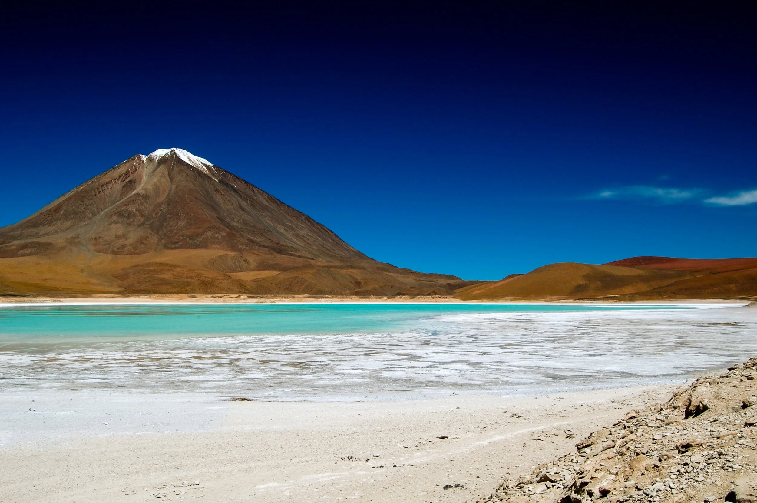 Donkere berg met sneeuwtop, heldere turquoise water, wit zoutvlaktes en droge, gele heuvels onder een blauwe hemel.