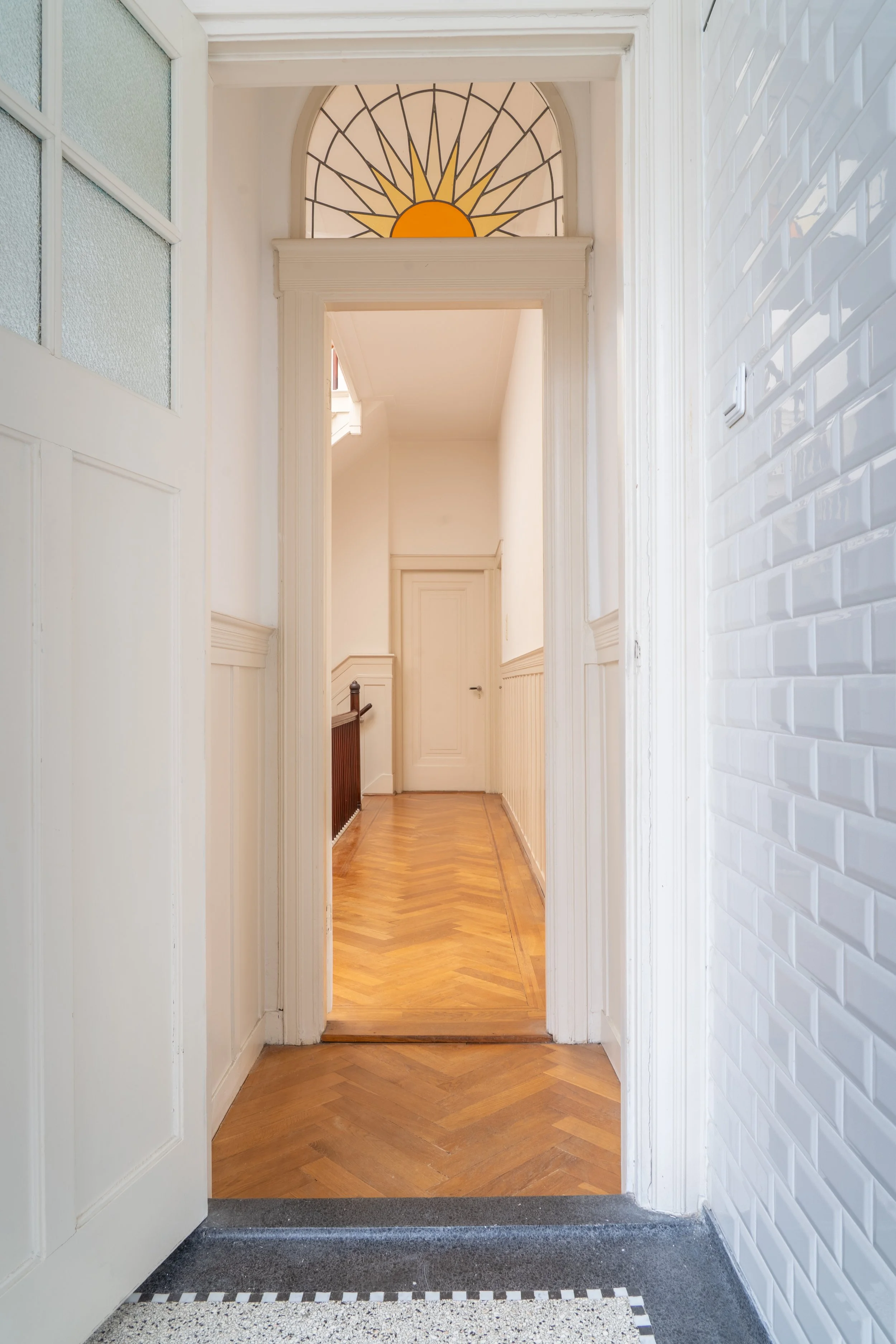 Binnenkomst met witte muren, een houten vloer en een doorlopende deur. Boven de deur is een raam met een geel/oranje zon en stralen, die de zon in een boog vorm tonen.