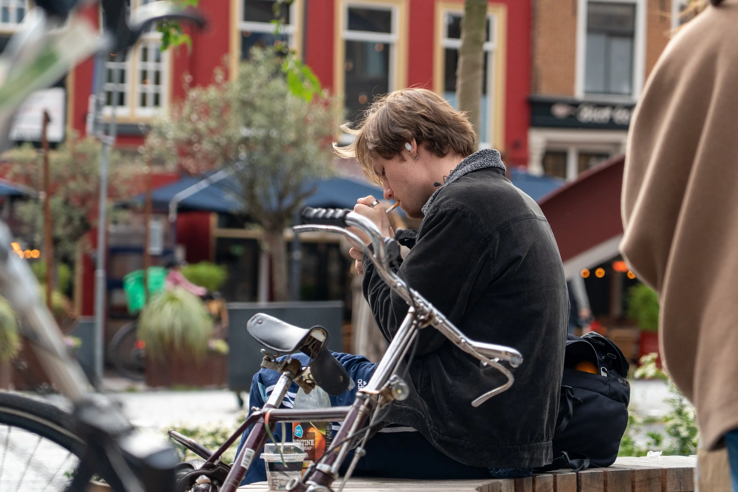 Jongeman met oorbel en oordopje zit op een bankje in een stadscentrum, terwijl hij rookt.