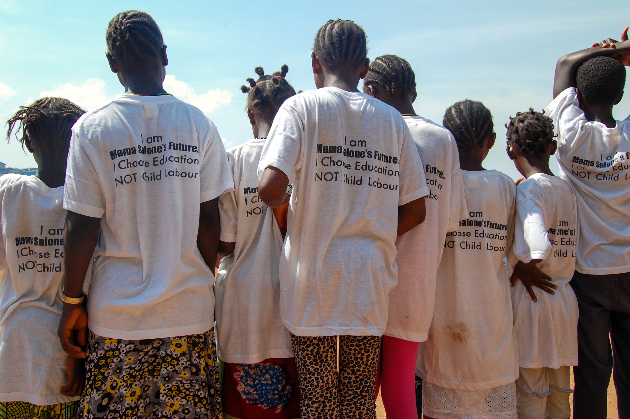 Groep jonge meisjes en vrouwen gezien van achter, dragen witte T-shirts met tekst: 'Ik ben Mama Salone's Toekomst. Ik kies onderwijs, geen kinderarbeid.'