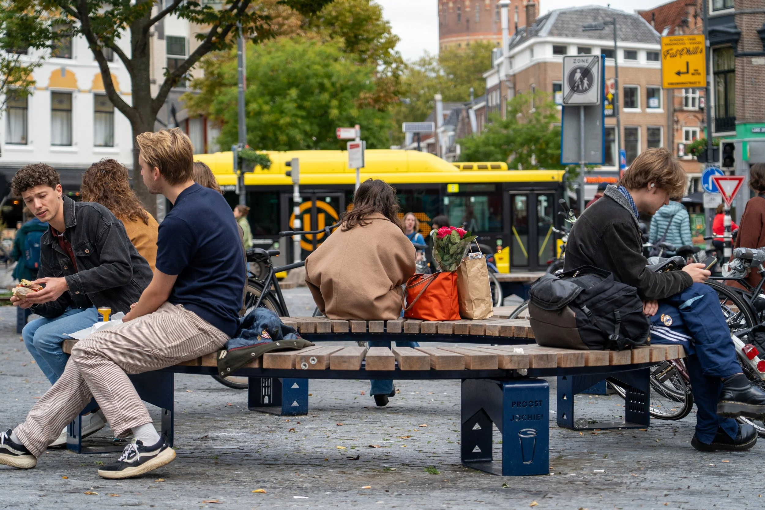 Mensen zitten op een bankje in een druk stedelijk plein, met een gele tram op de achtergrond, bomen en gebouwen. Sommigen eten, anderen kijken op hun telefoons.