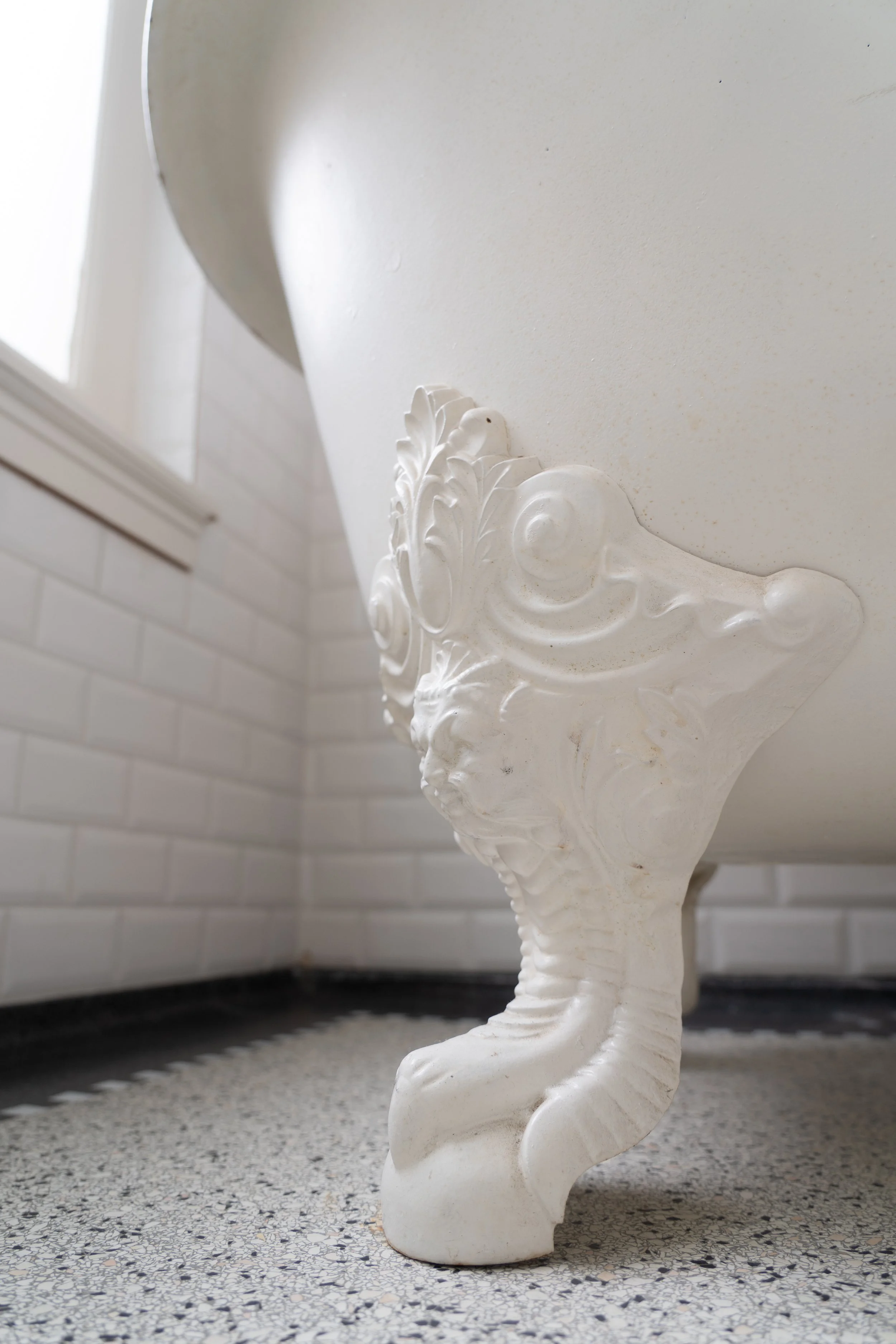 Een wit, sierlijk toilet met een gedetailleerd versierd verwijderbaar deksel, geplaatst op een vloer met een kleine, stoeptegelfestijn. De achtergrond toont een wit betegelde muur en een raam dat licht binnenlaat.