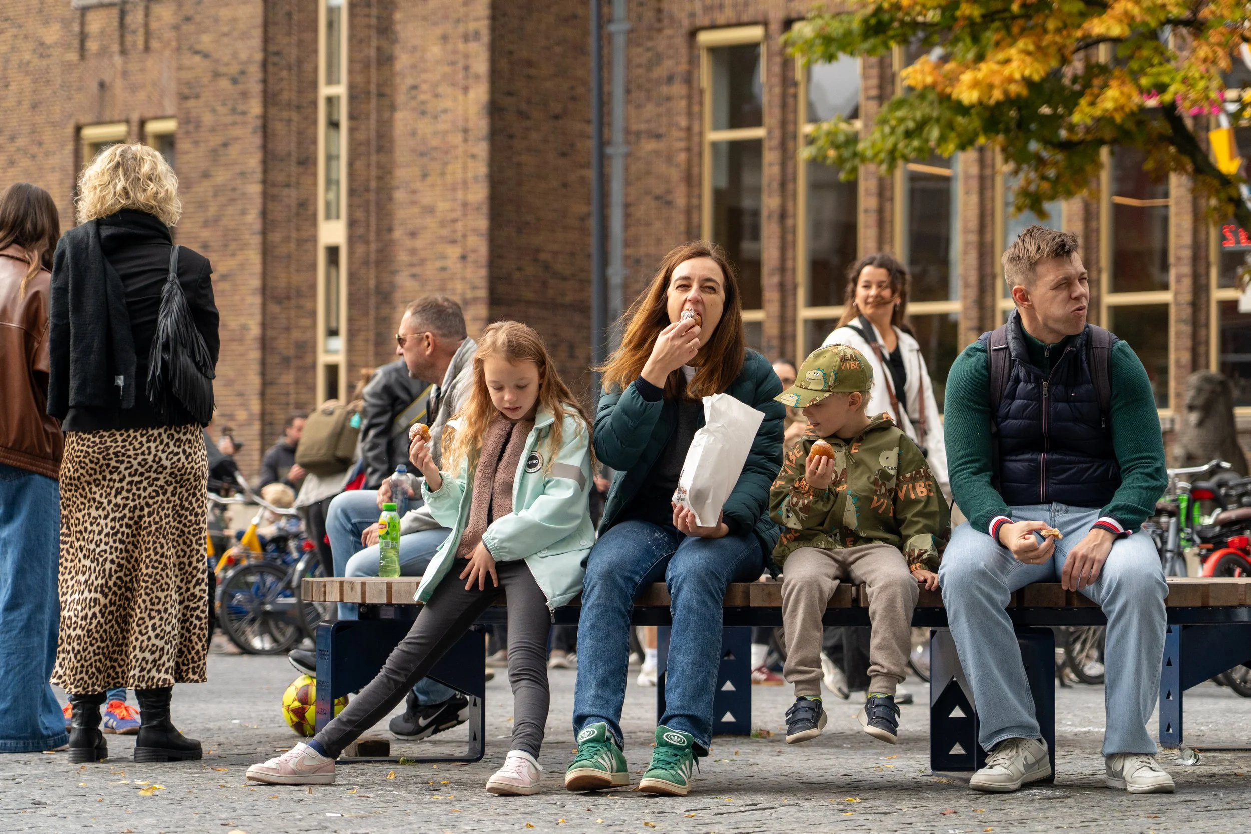 Mensen zitten op een bankje in een stedelijke omgeving, genietend van snacks en gesprekken, met fietsen en een boom met herfstbladeren op de achtergrond.