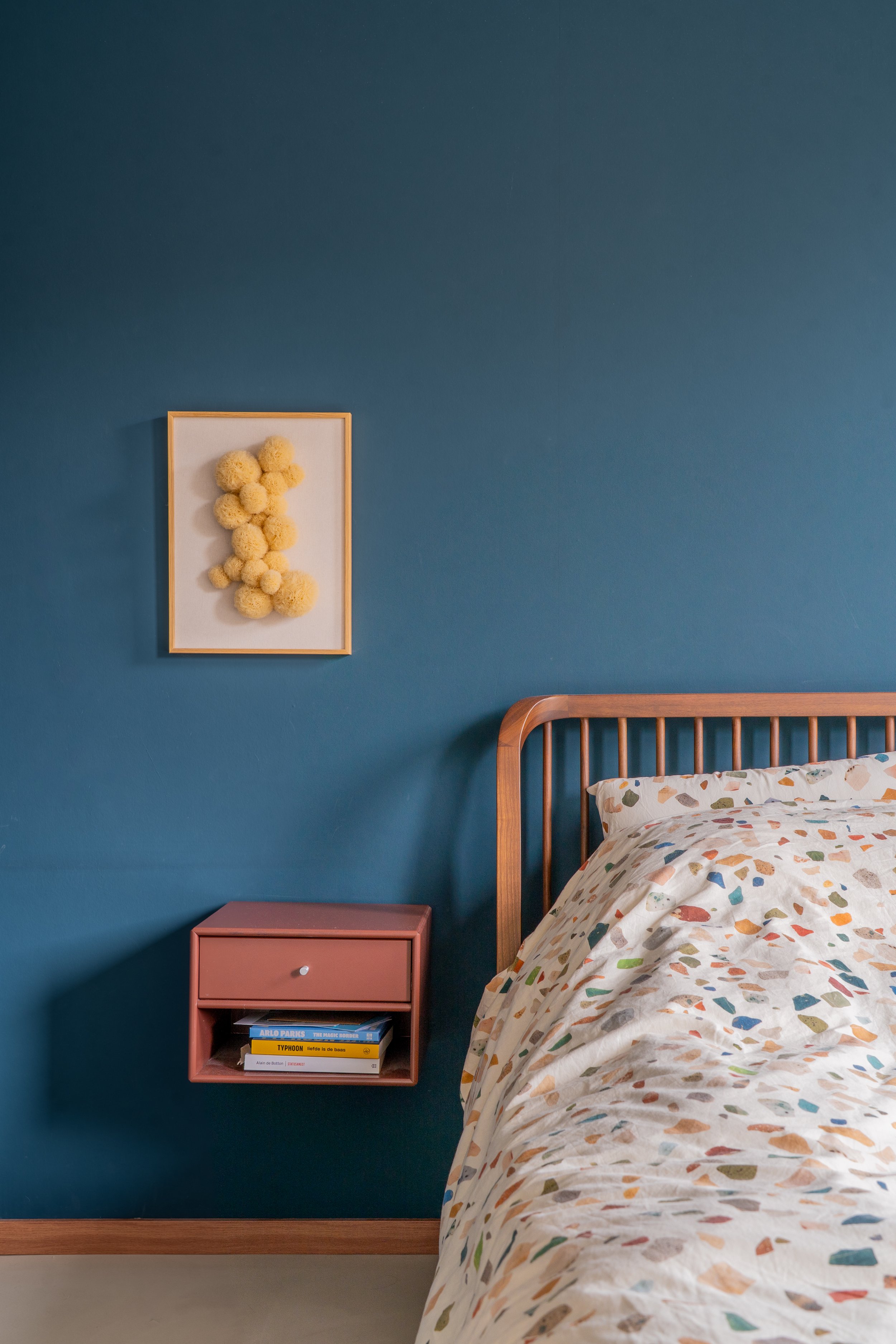 Een slaapkamer met een blauw muur, een afgeronde houten bedframe, een bed met een beddengoed met gekleurde blob-prints, een gekleurde nachtkastje met boeken en een aards kleurige muurdecoratie van pompons