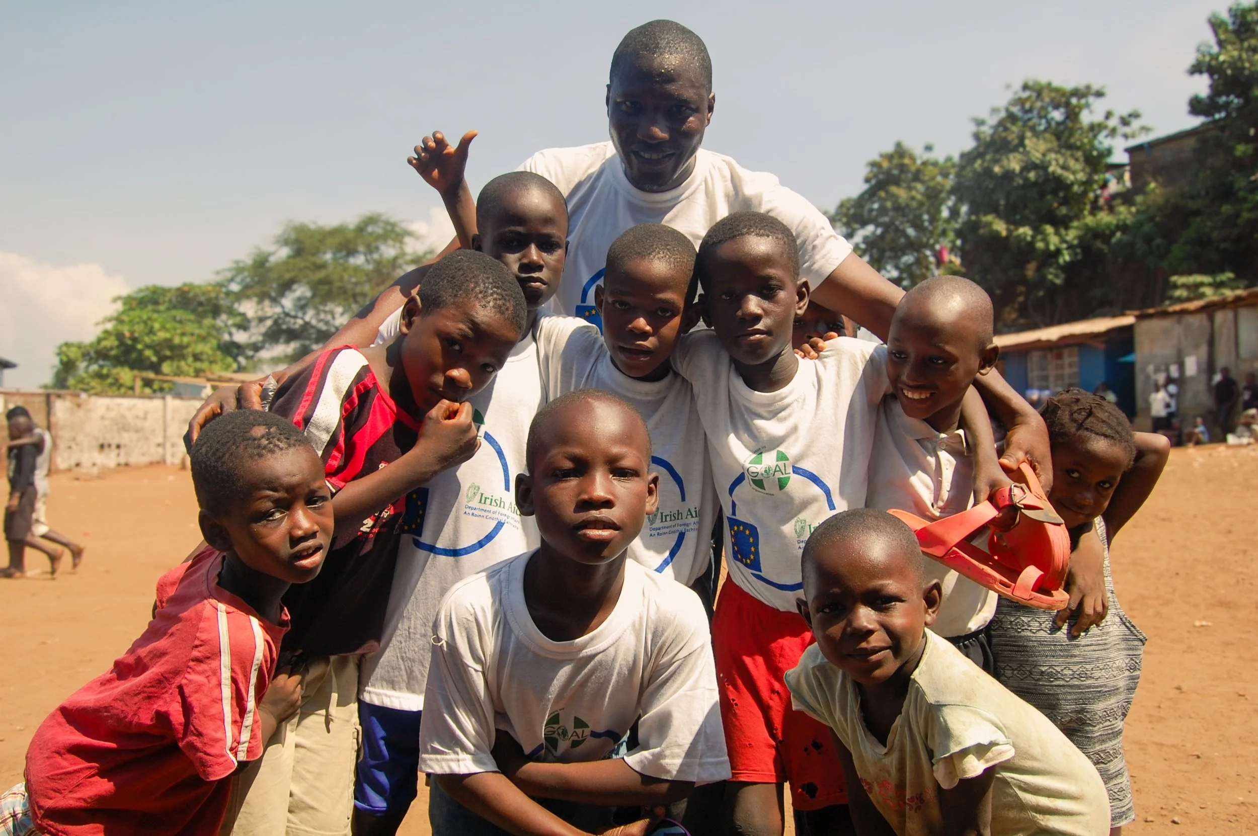 Group of children and a man outdoors on a sunny day, with some trees and buildings in the background.