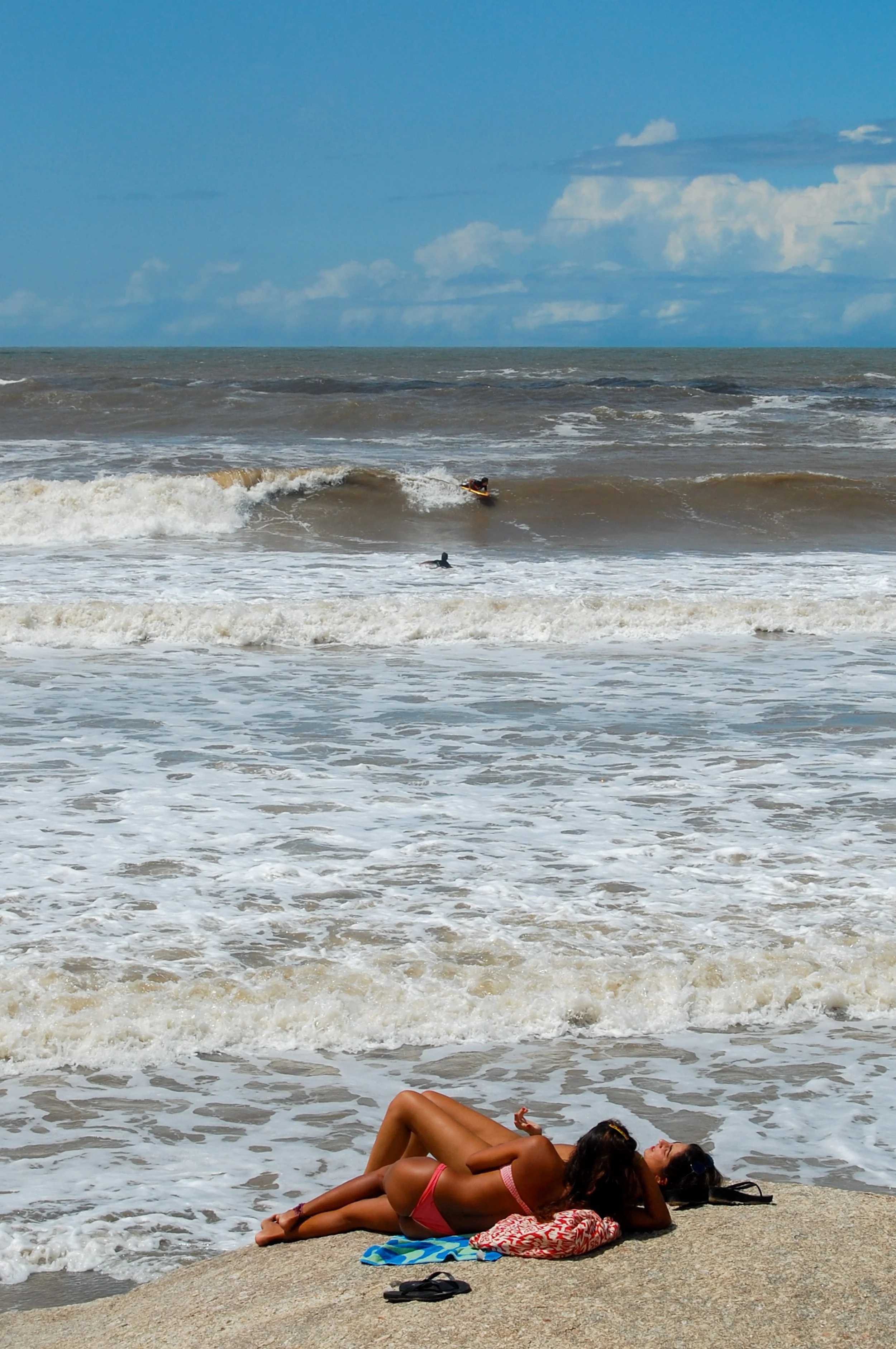 Vrouw in een roze bikini ligt op een handdoek op het strand, terwijl ze naar de zee kijkt. De achtergrond toont golven en een bewolkte hemel.