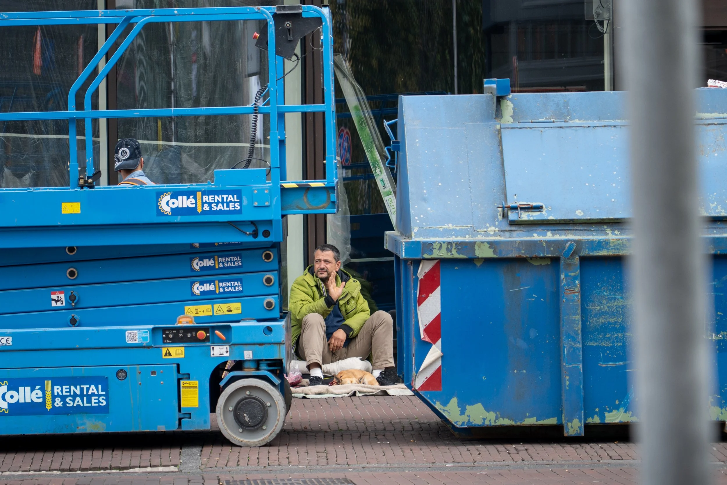 Twee mannen zitten onder een hijskraan die is voorzien van een blauwe hijskraan, met een hond die rust op een deken. Een man in een groene jas zit naast de hond en praat met de andere die in een blauw uniform staat. Op de achtergrond een glazen gebou