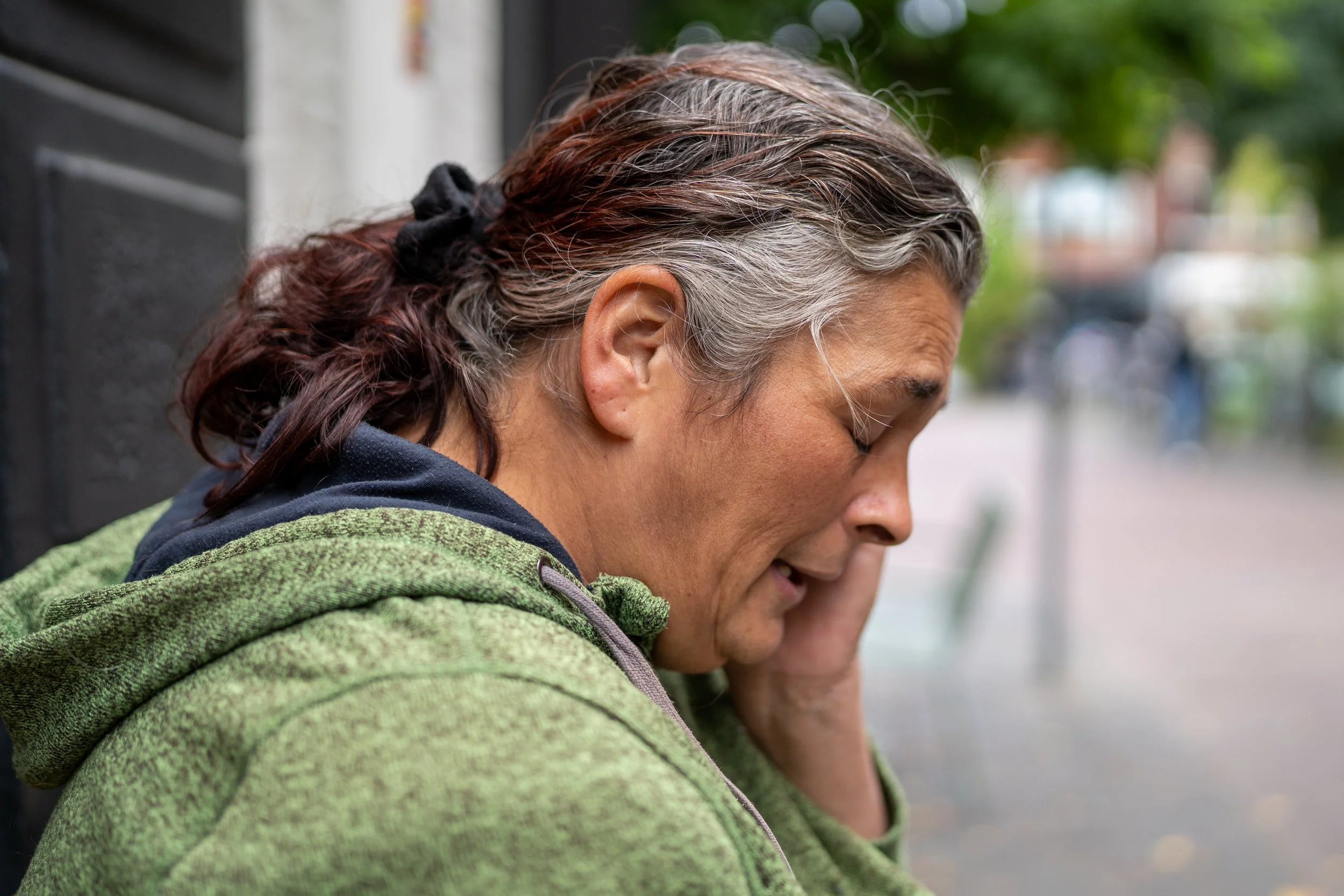 Close-up van een vrouw met grijs en bruin haar in een paardenstaart, gekleed in een groene jas, let op haar gezicht en haar oor, op een straat met groenbomen op de achtergrond.