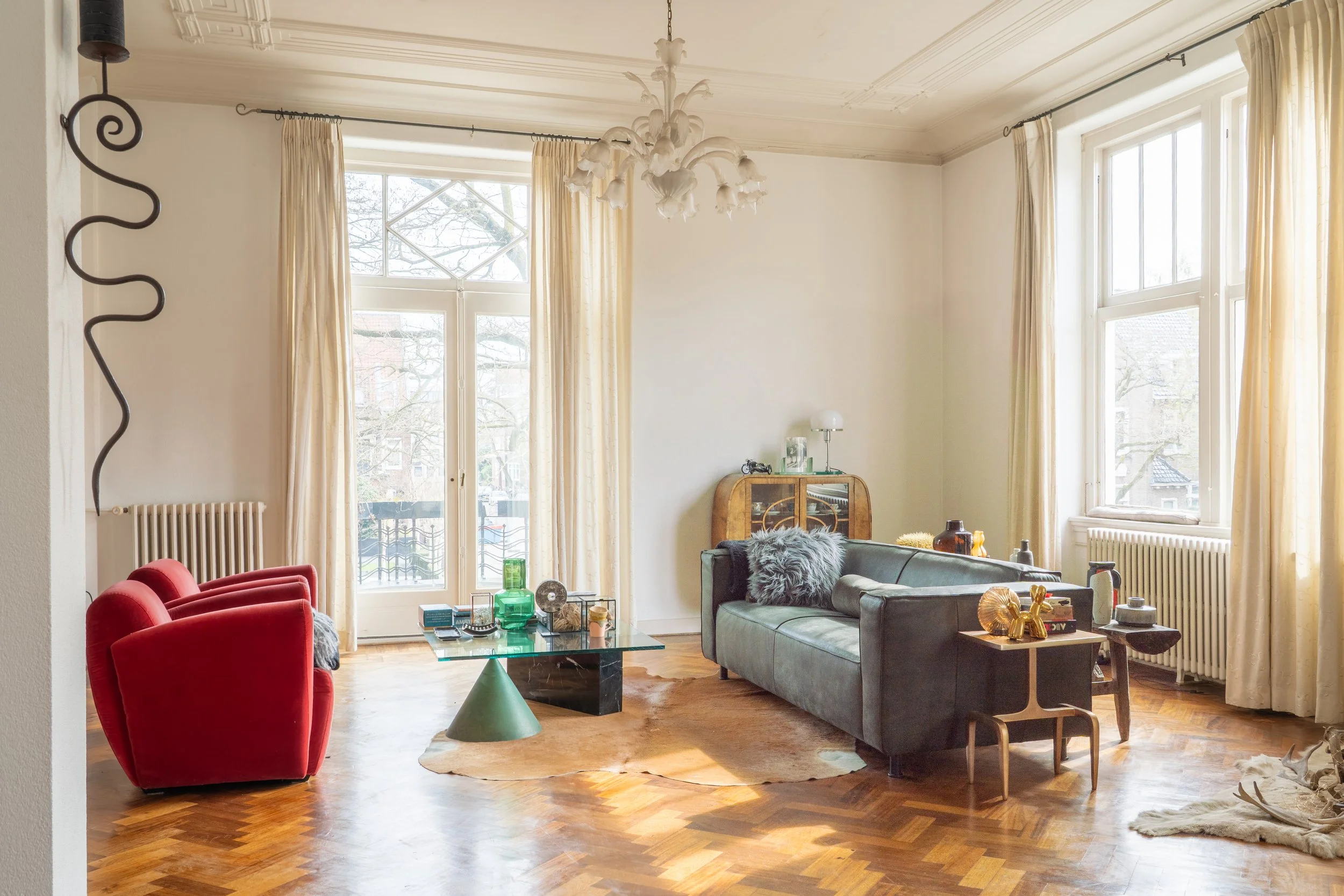 Woonkamer met grote ramen, goudgele gordijnen, een kristallen chandelier, groene en rode stoelen, een glazen salontafel, en decoratieve spullen op een tafel en plank.