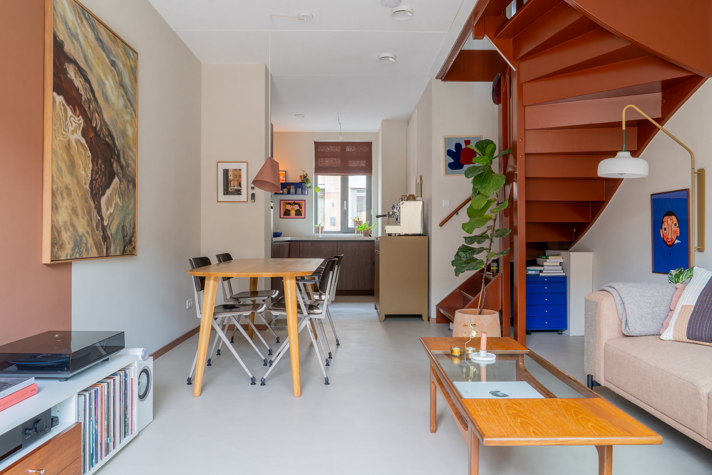 Interieur van een moderne woonkamer en eetkamer met een houten eettafel en zwarte stoelen, een beige bank met kussens, kunstwerken aan de muur, een houten boekenplank en een trap met houten treden en rode metalen ombouw.