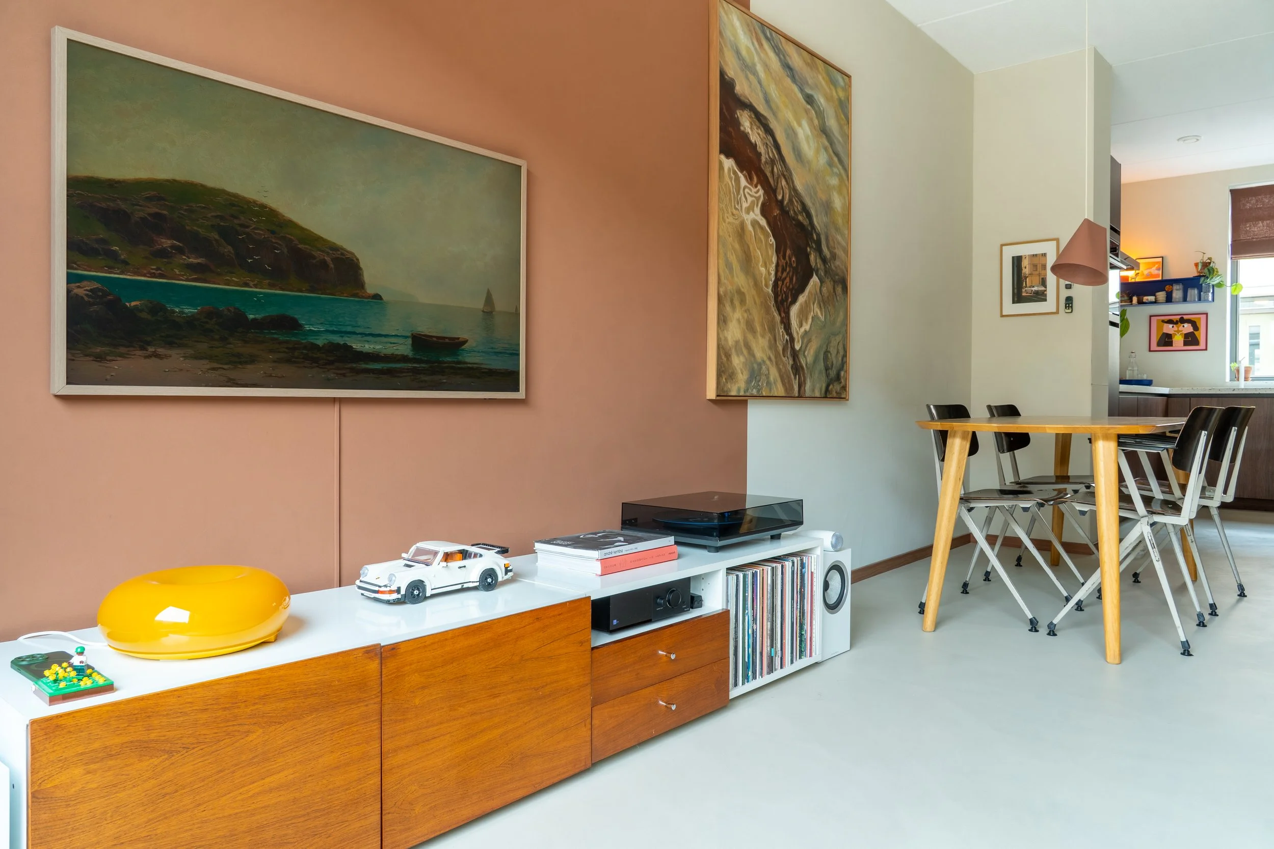 Leefruimte met wanddecoraties, een witte entertainmentkast met speelgoed, vinylplaten en elektronische apparaten, een hoge eettafel met zwarte stoelen, en een keuken op de achtergrond.