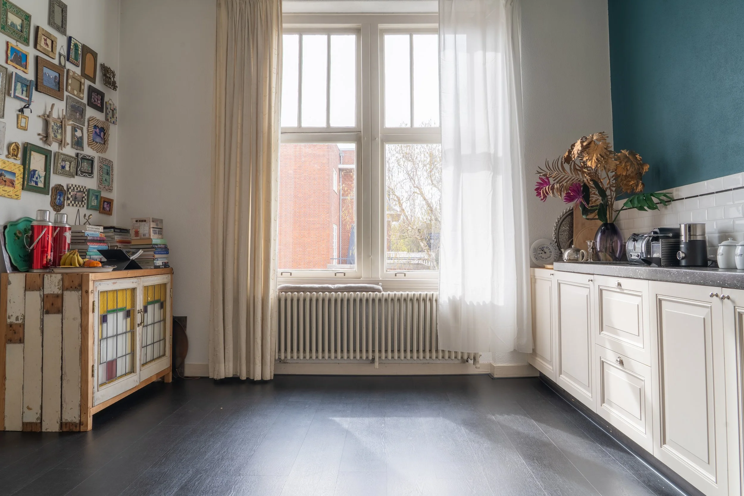 Een keuken met witte kasten, een grote raam met witte gordijnen, een blauwe muur, een vaas met bloemen, een kookplaat, en een retrokast met boeken en fruit.