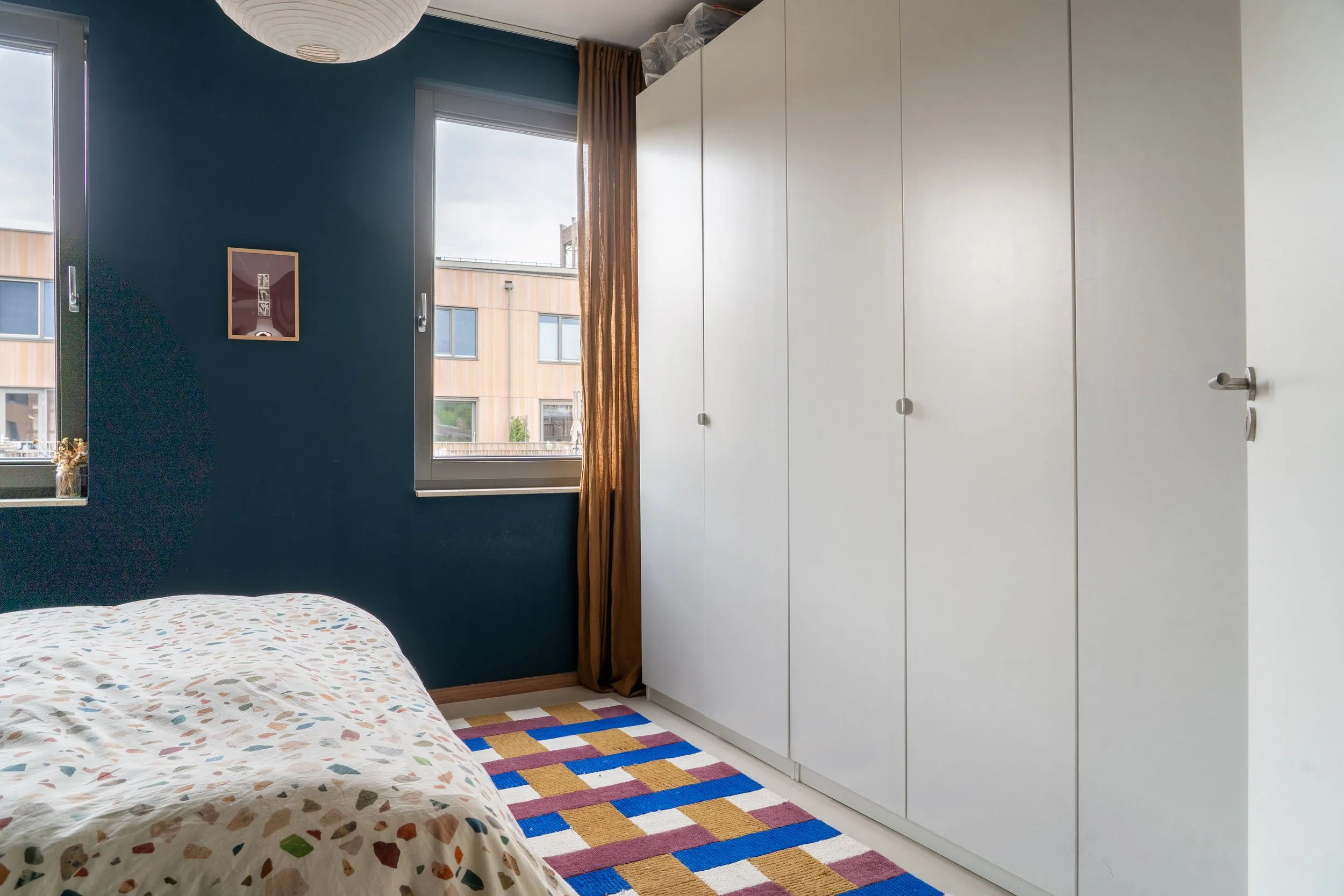 Een slaapkamer met een bed, een grote witte kast, twee ramen en een muur die donkerblauw is. Er hangt een witte lampenkap aan het plafond en er ligt een kleurrijk vloerkleed op de vloer.