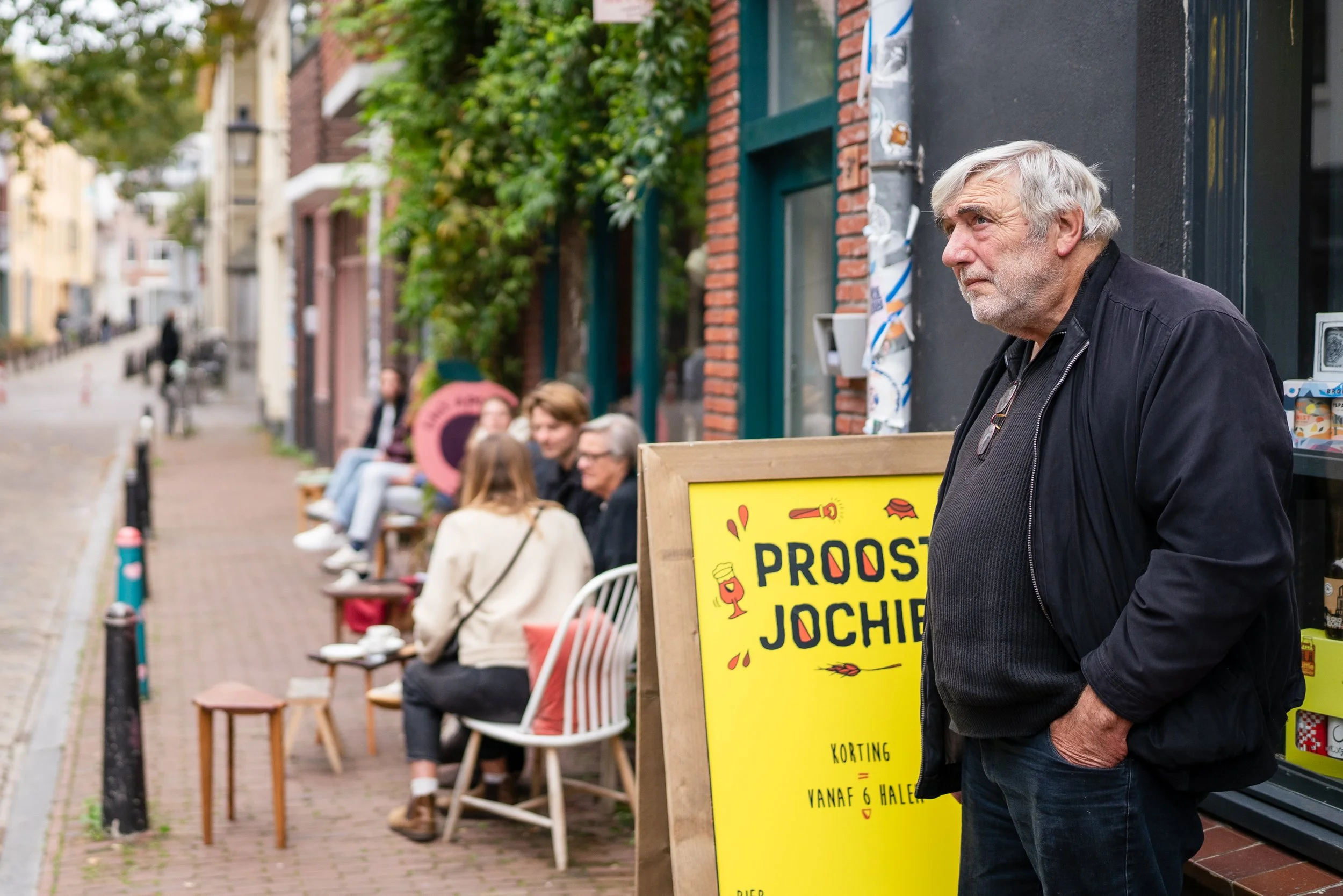 Een oudere man met grijs haar en een zwart jack staat naast een gele prijslijst buiten een café, terwijl mensen op een terras op de achtergrond zitten.