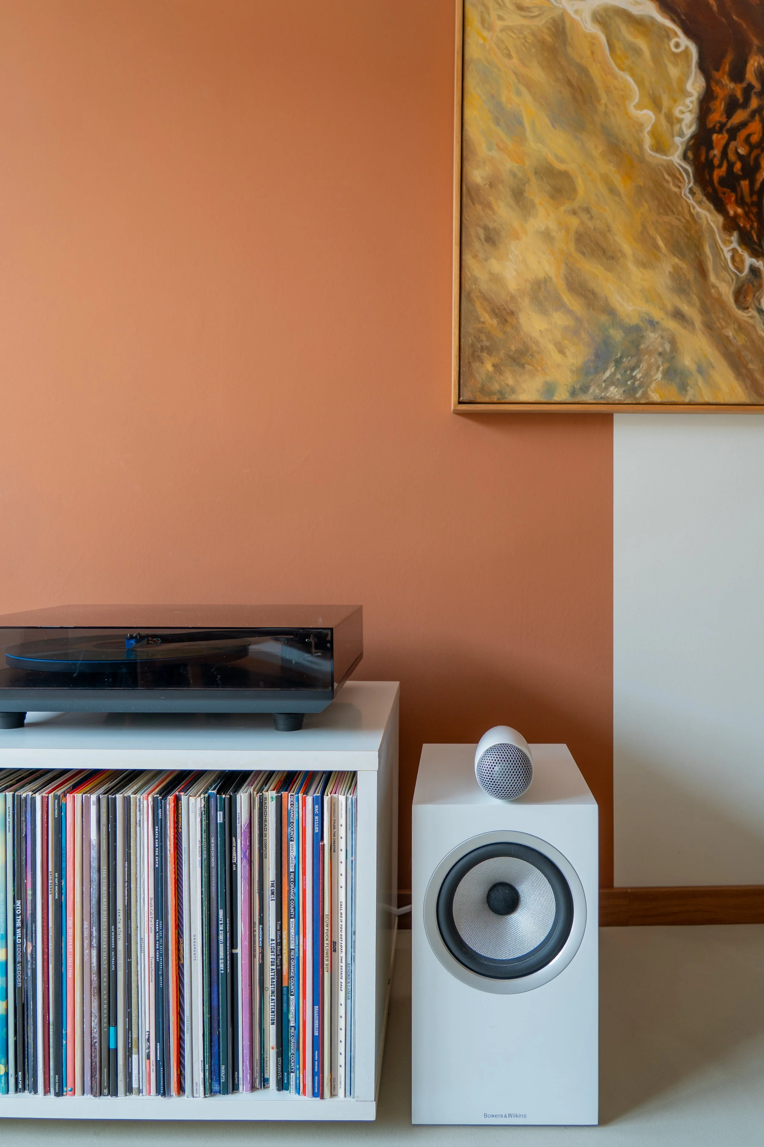Een platenspeler op een witte kast met een grote collectie vinylplaten eronder, een witte luidspreker op een naastgelegen wit apparaat, een schilderij aan de muur en een muur in twee kleuren (roze en wit).