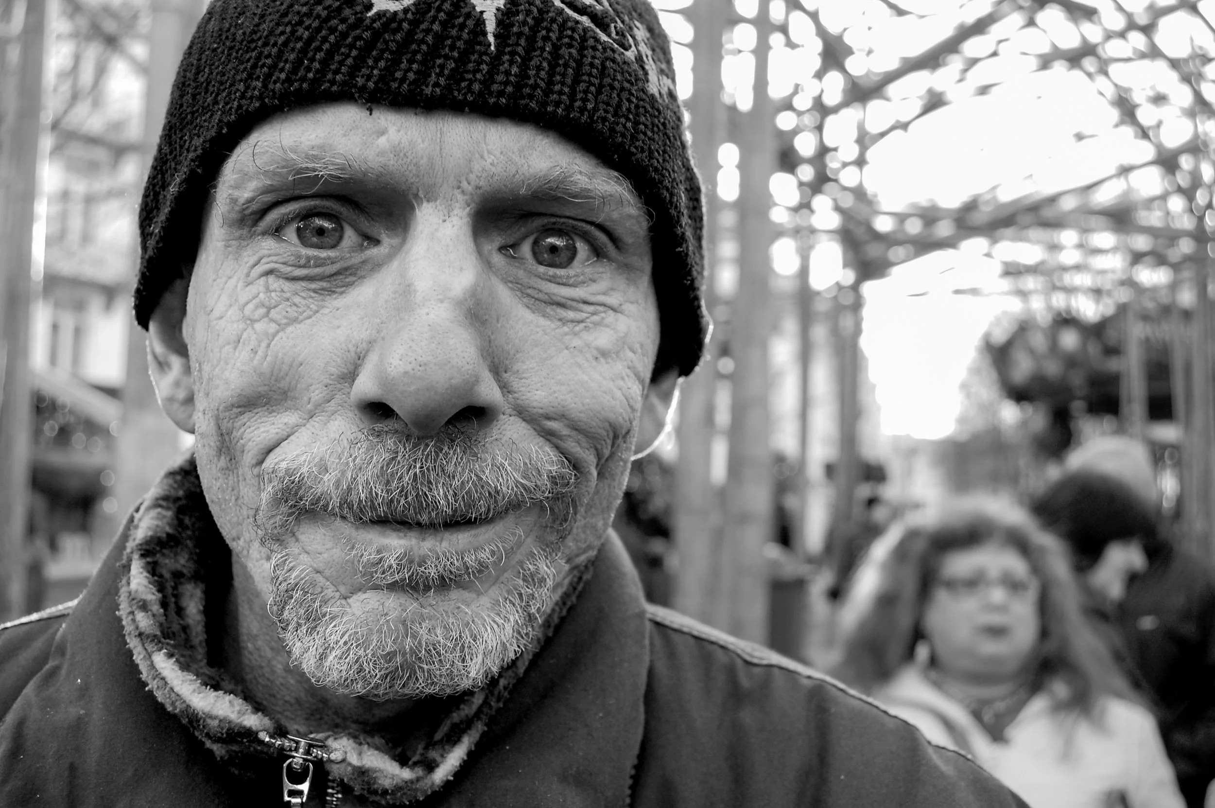 Close-up van een oudere man met grijze baard en wenkbrauwen, zwart petje en jas, op een gespitst, bokeh-achtergrond in een park of bos, zwarte en witte foto.