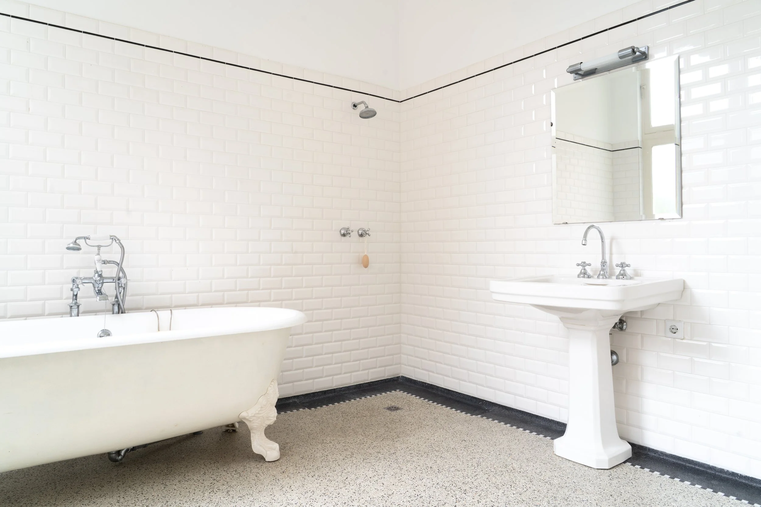 Een wit vintage bad met poten, naast een wit wastafel met twee kranen, een spiegel en een radiator. De muren zijn bedekt met witte bakstenen tegels met twee dunne zwarte lijnen.