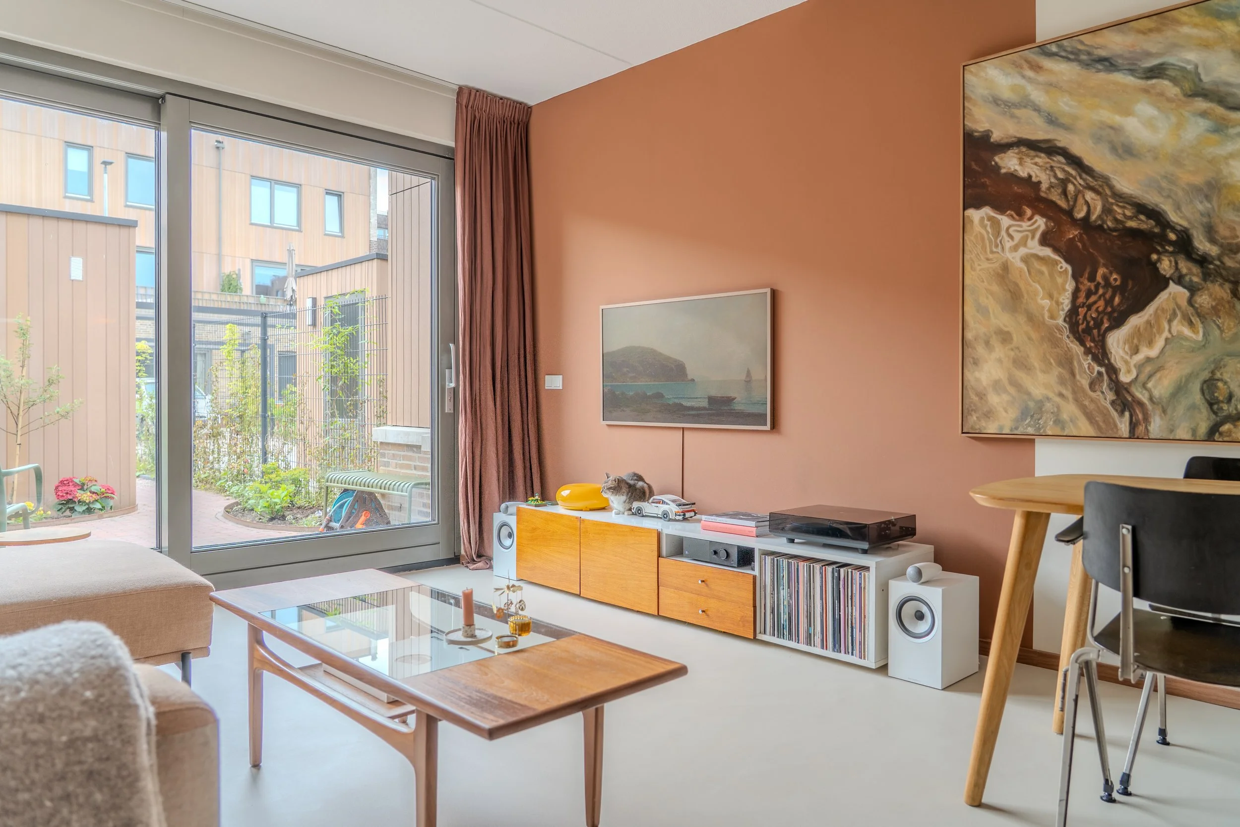 Een woonkamer met grote glazen deuren die naar een tuin leiden, met een satijnen roze muur, muurkunst, en een houten meubel met modellenauto's en een luidspreker. Er zitten katten op de meubel.