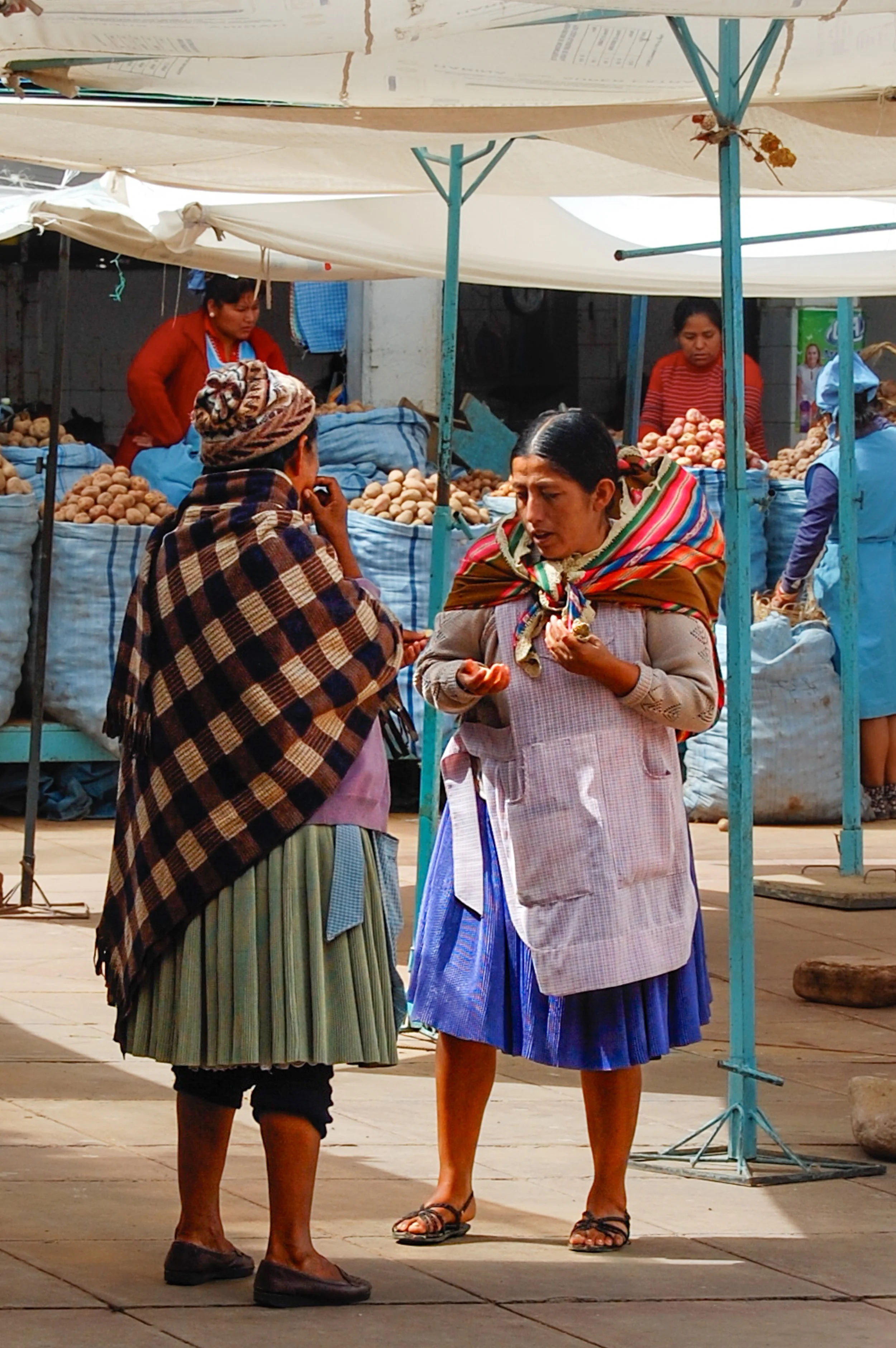 Een markt met drie vrouwen die gesprekken voeren. Twee vrouwen in de voorgrond dragen traditionele kleding en lijken in een gesprek verwikkeld. Op de achtergrond staan meer mensen en kramen met aardappels. Ze bevinden zich onder een tent met blauwe e