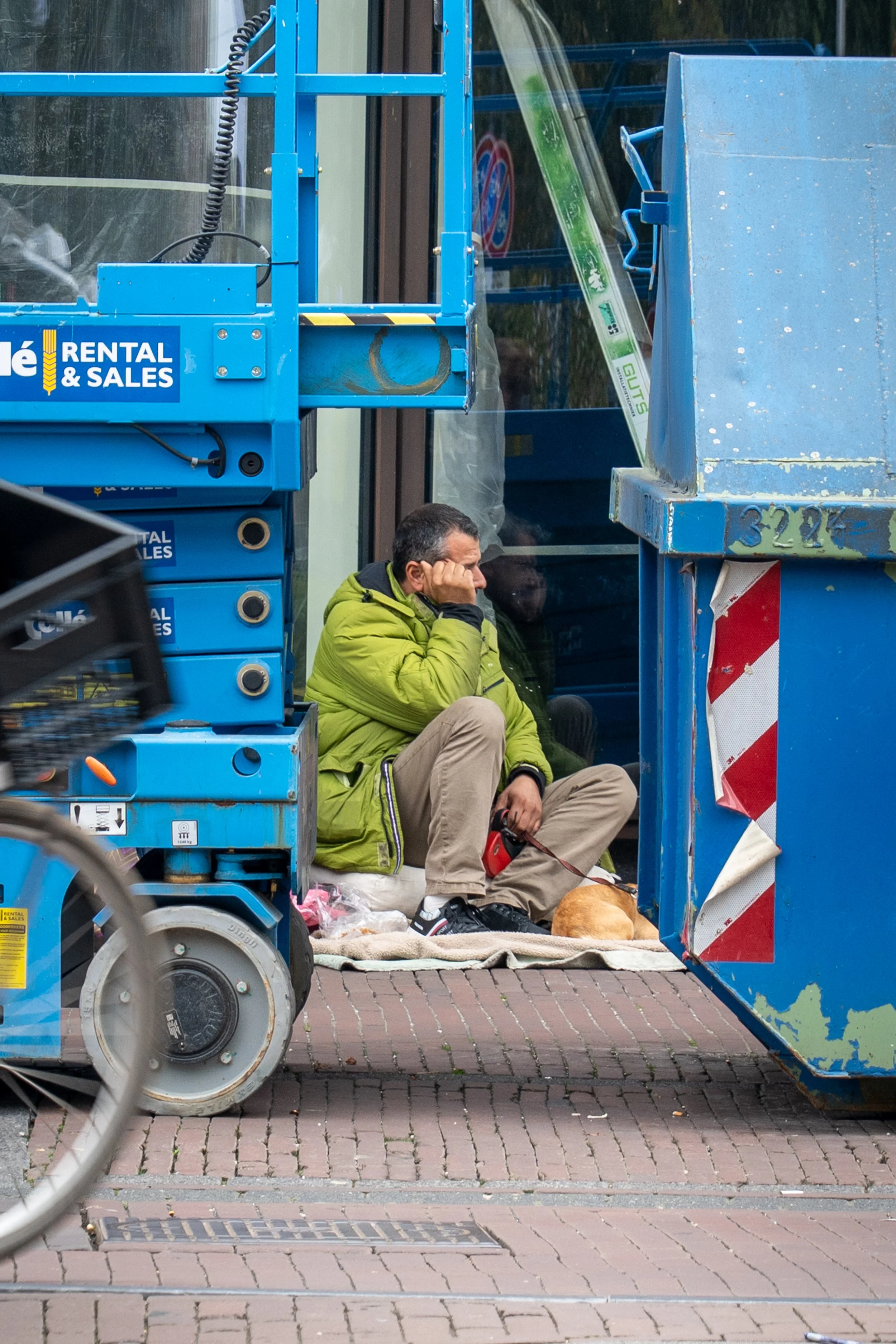 Man in een groene jas zit op de grond naast een dakloze met een hond, tussen bouwmaterialen en een gereedschapswagen, op een straat met bakstenen.