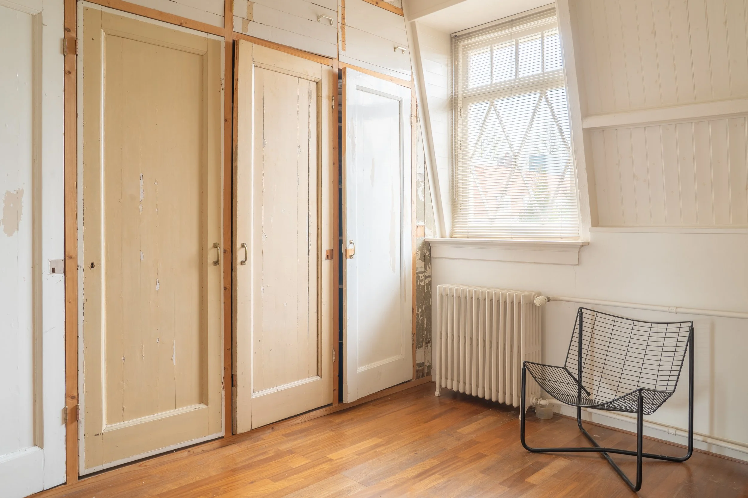 Een kamer met houten vloer en witte gestuukte wanden. Een grote raampartij met open jaloezieën, een radiator en een zwart metalen stoel. Een muur met kastdeuren in aanbouw.