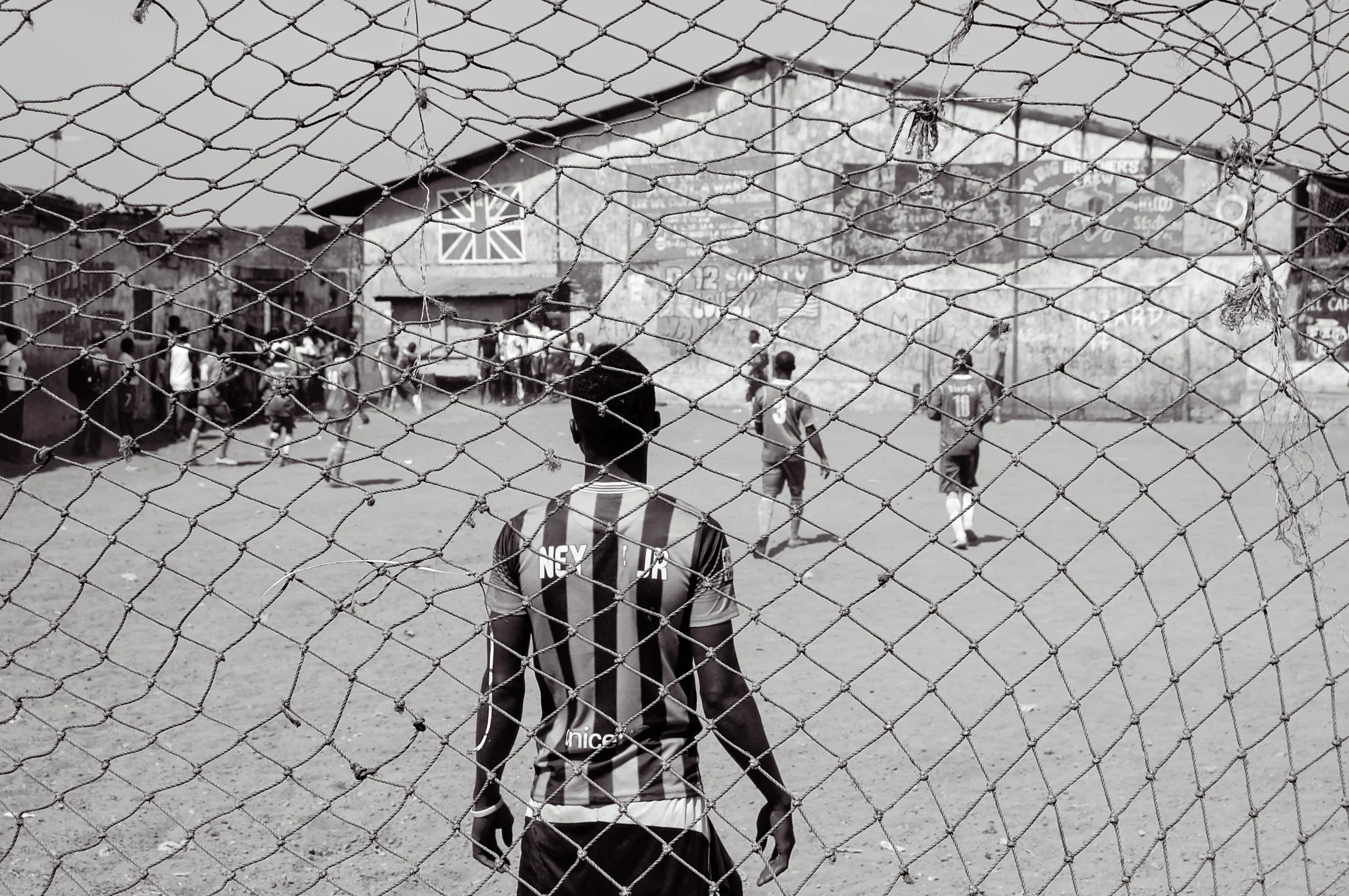 Zwart-wit foto van een voetbalveld met kinderen die spelen, gezien door een afscheidingshek.