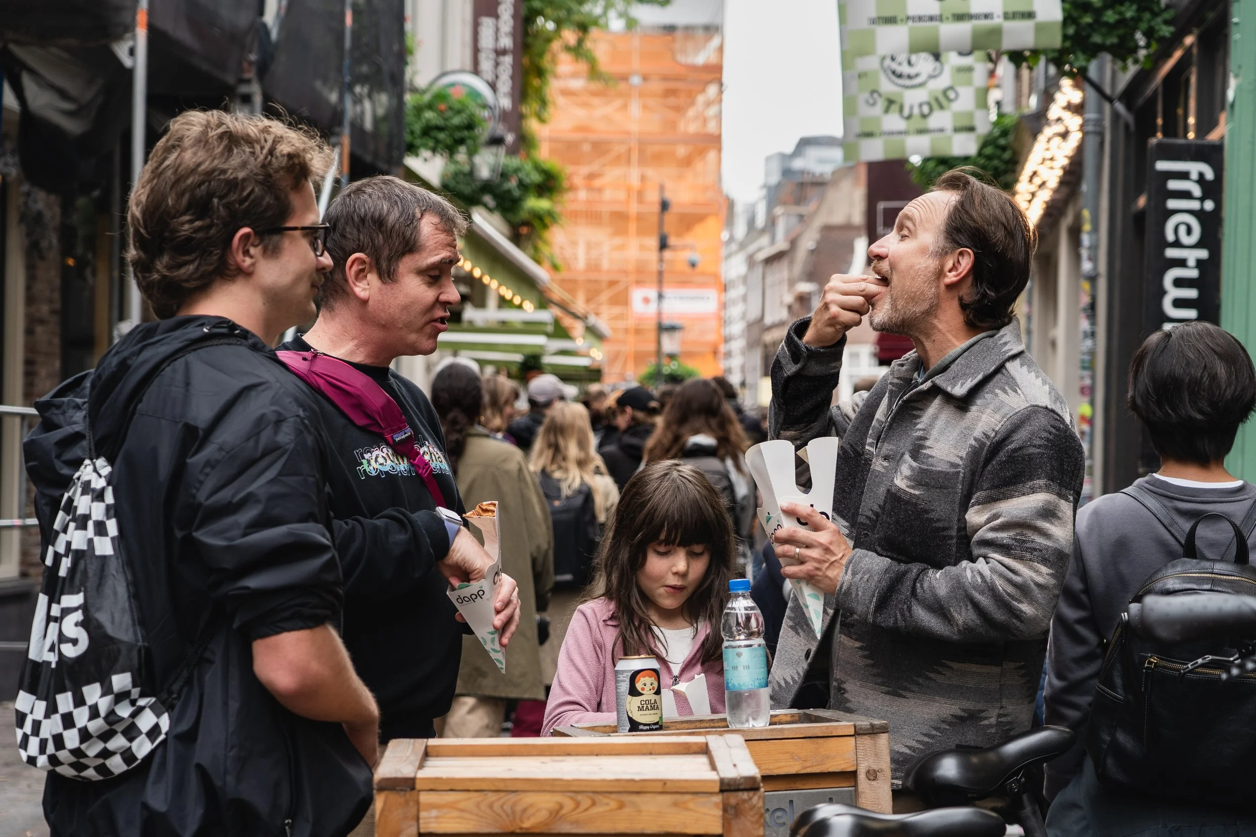 Mensen in een straatmarkt of evenement, met drie mannen en twee kinderen die eten en drinken met winkels en bouwactiviteiten op de achtergrond.