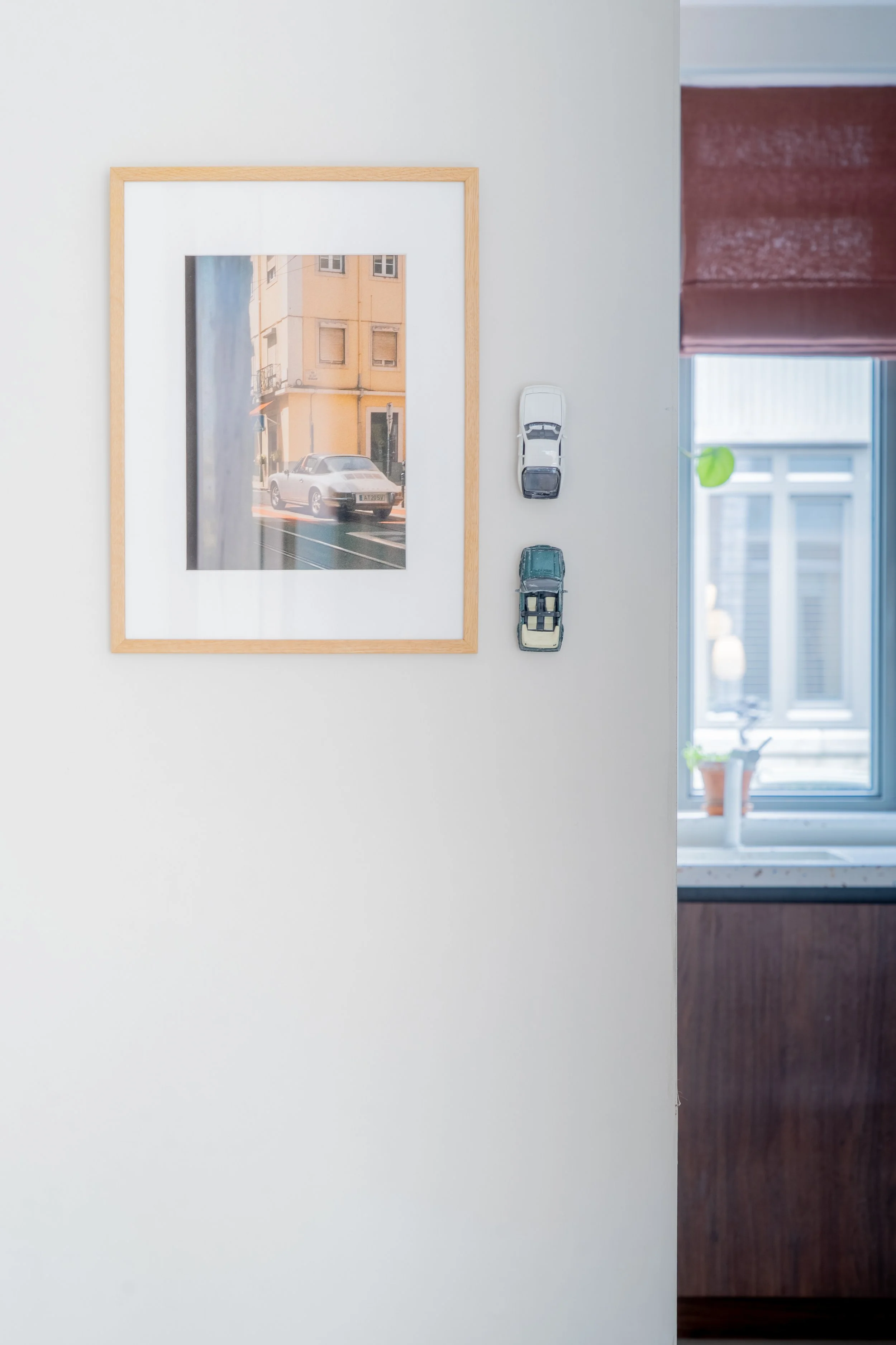 Binnenzicht van een kamer met een muur met een foto van een straat met een witte sportwagen, twee kleine modelauto's hangen aan de muur, en een raam met een rood gordijn en een plantenpot op een vensterbank.