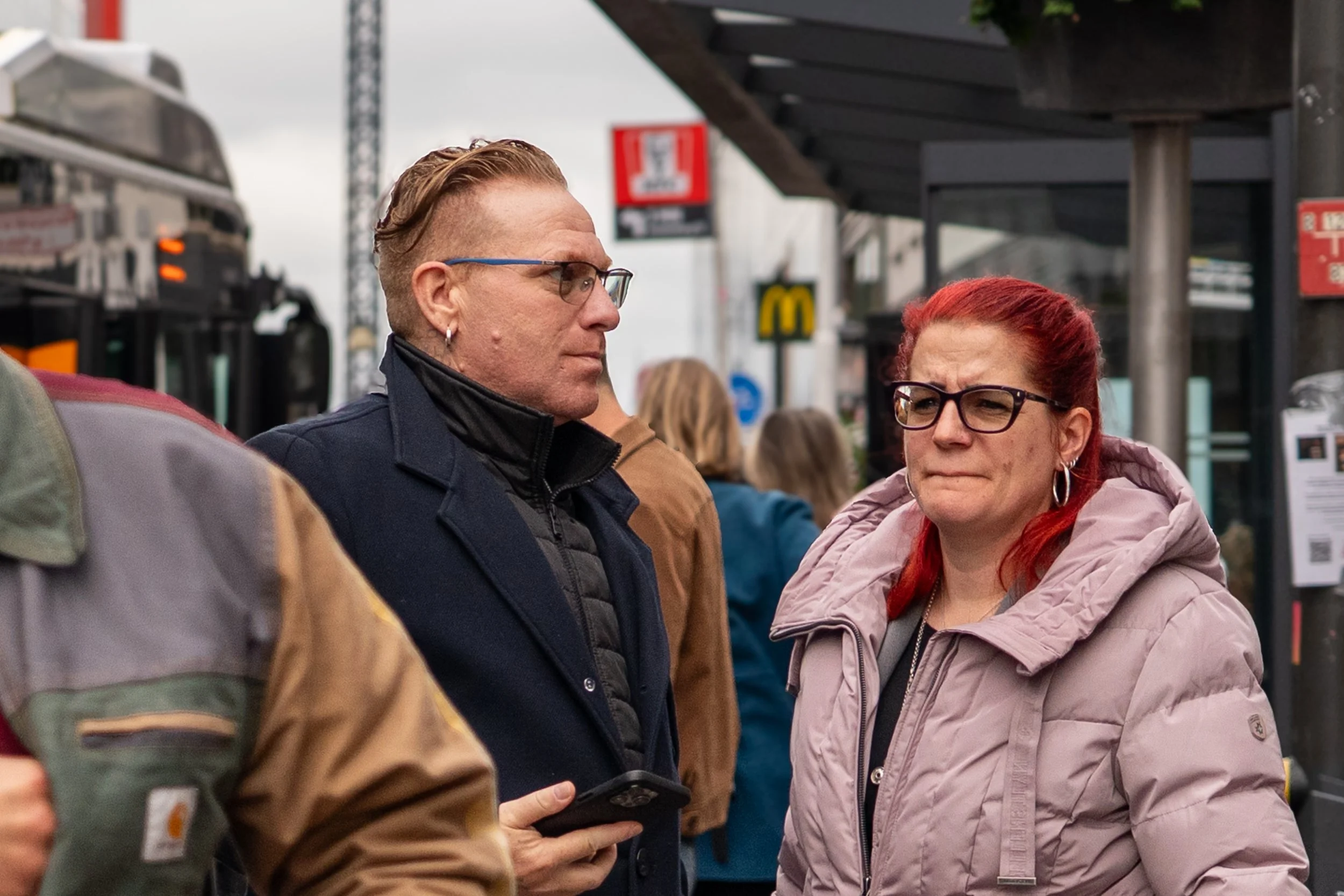 Twee mensen, een man met bril en een vrouw met rode haar en bril, staan in een drukke omgeving buiten, mogelijk op een markt of station.