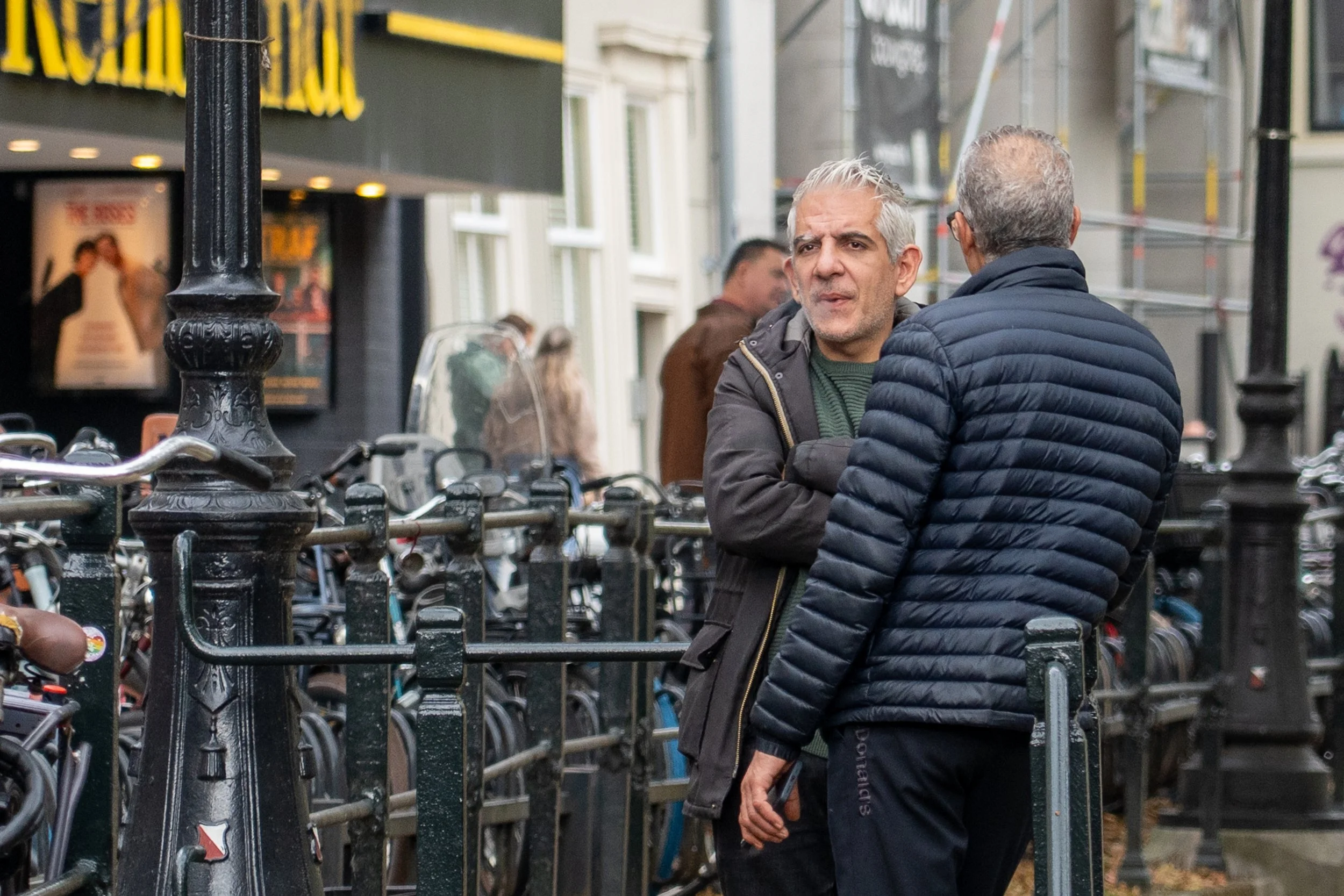 Twee oudere mannen praten op straat, omgeven door fietsen en een zwarte metalen hekwerk.