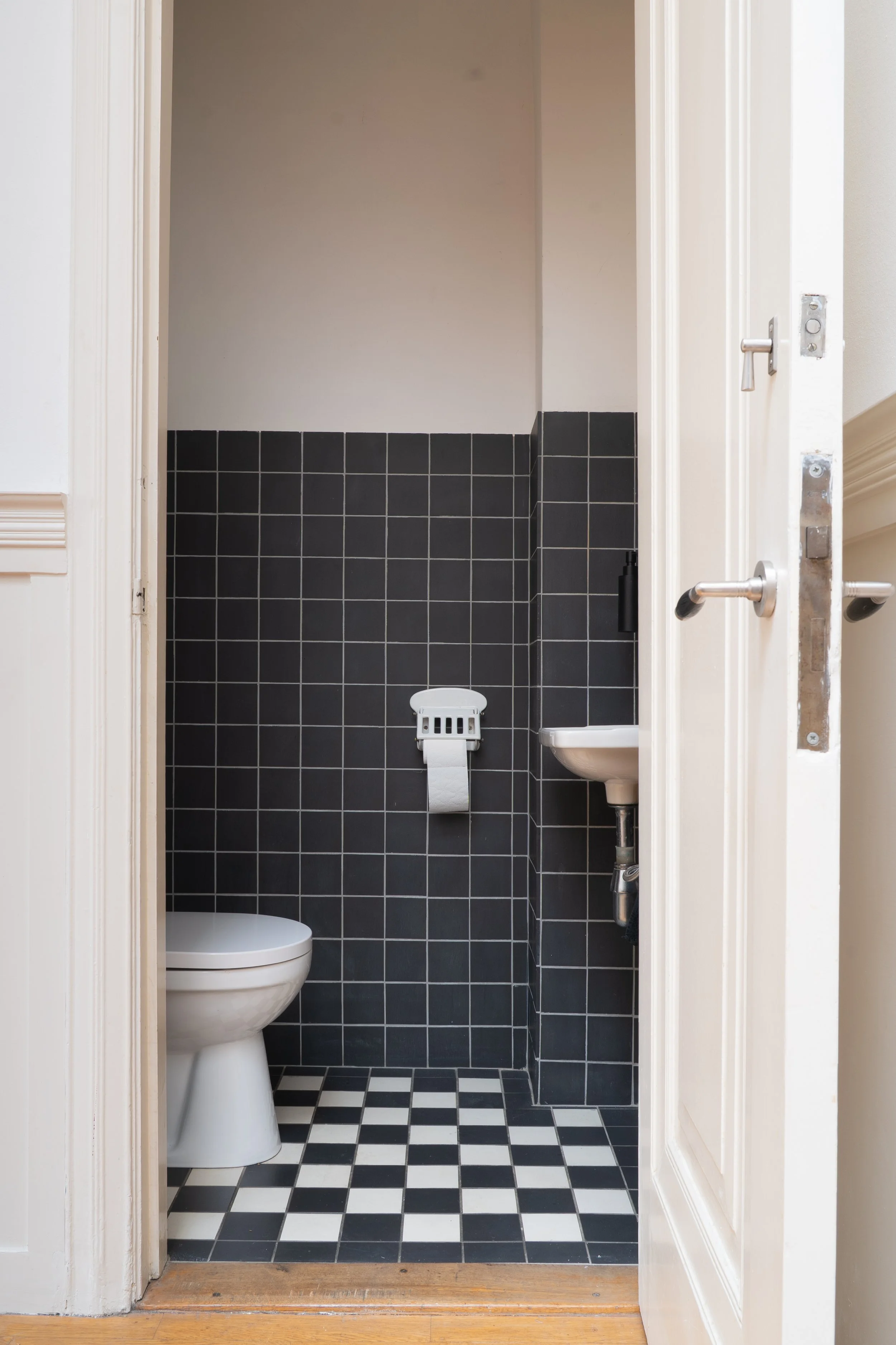 Kleine toiletruimte met zwarte tegels, een wit toilet en een klein lavabo, zicht vanaf de deuropening met een houten vloer.