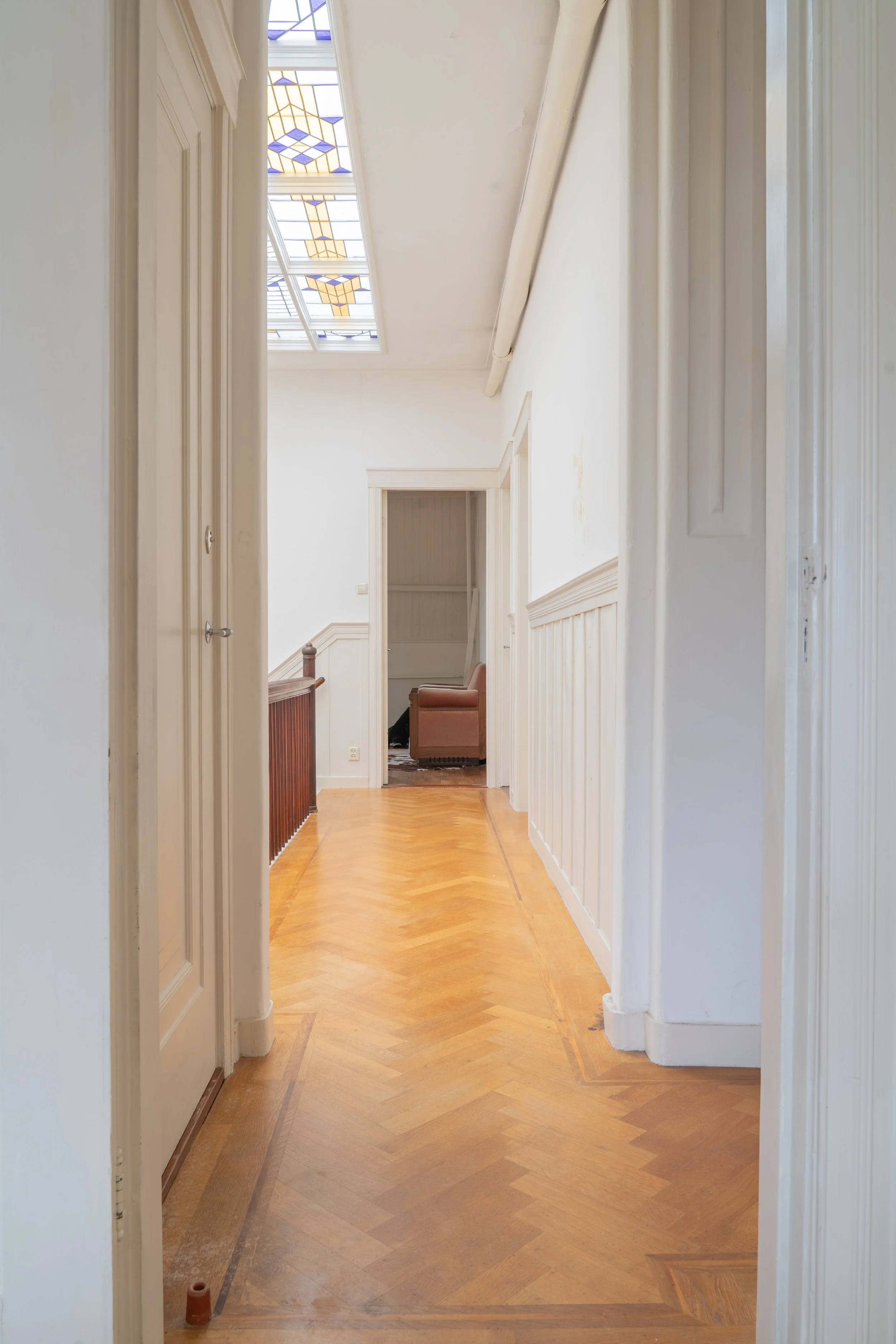 Een lange, smalle gang met houten vloer, witte muren en plafond, en een plafondraam met gekleurd glasstalen raampanelen dat licht binnenlaat.