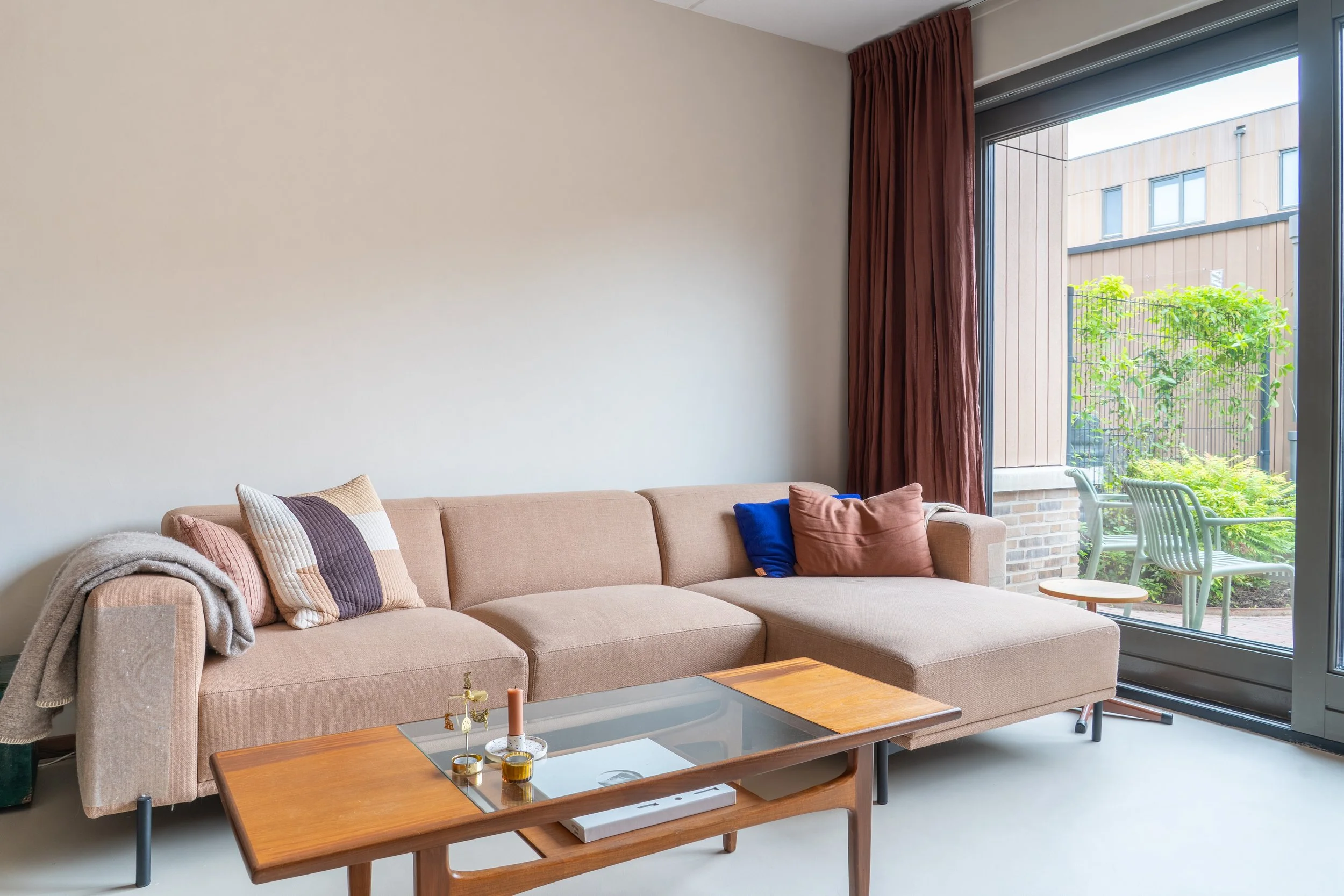 Een woonkamer met een lichtroze bank en verschillende kussens, een houten salontafel met decoraties en een grote raam met uitzicht op een terras en groene planten.
