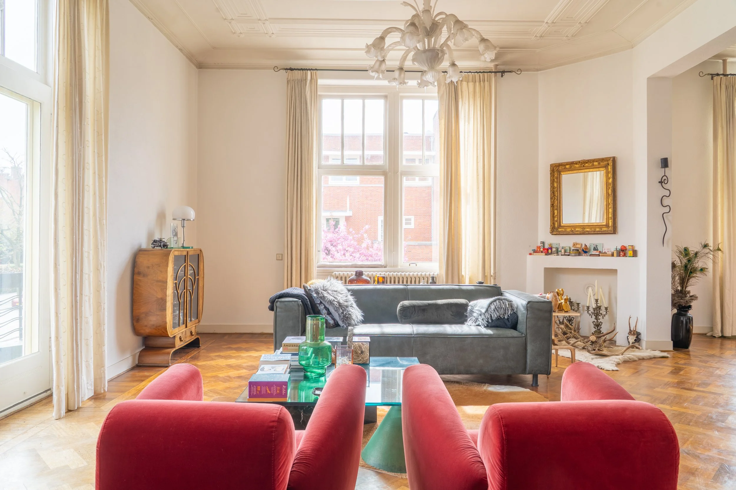Een woonkamer met grote ramen, een grijze bank, een koffietafel met groene decoraties, een spiegel in een gouden raam, en diverse decoratieve items. Er zijn rood gekleurde stoelen voor de koffie tafel en de vloer is van hout.