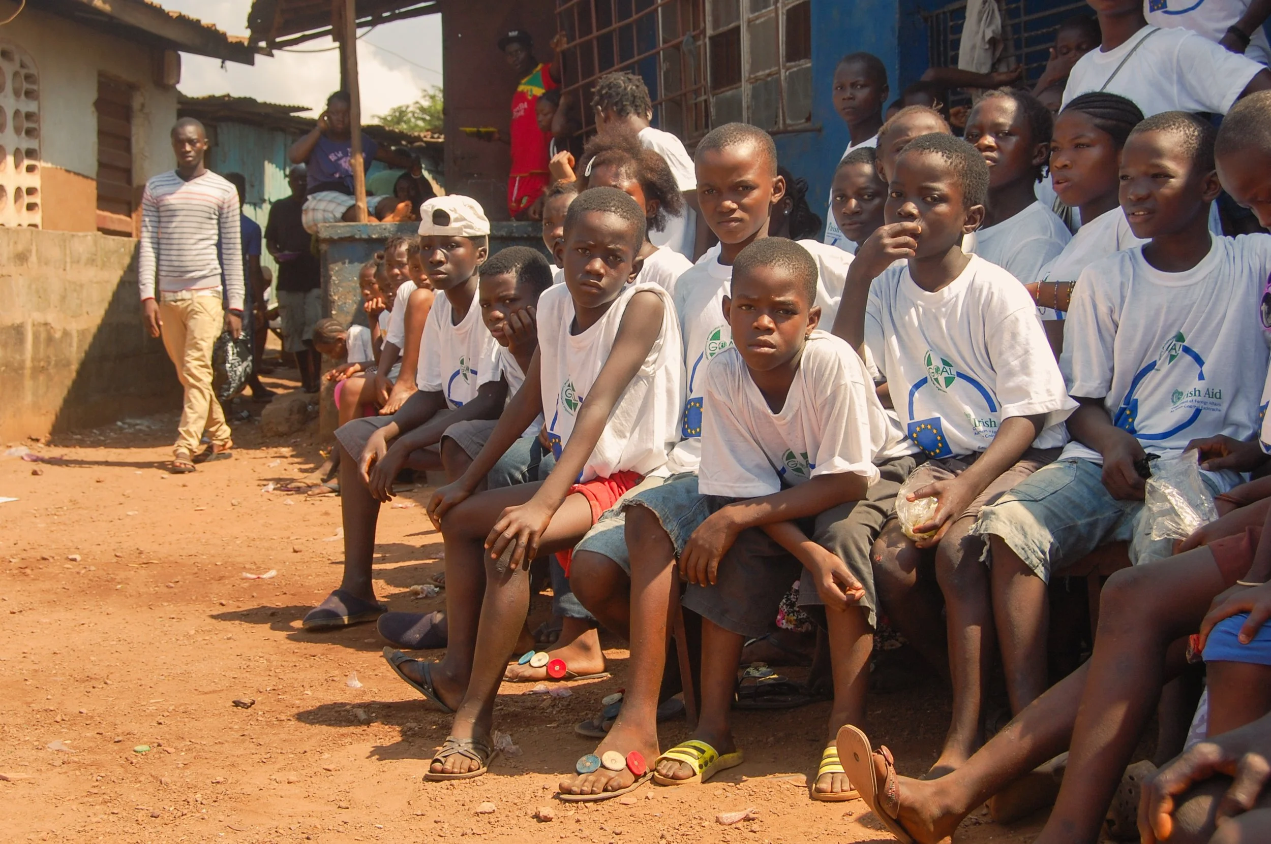 Een groep kinderen zit buiten op een rand in een gemeenschap, waarvan sommige kinderen zitten en anderen staan. Ze dragen zomerkleding en zien er bezorgd uit. Achter hen staan eenvoudige huisjes en een man loopt in de achtergrond.