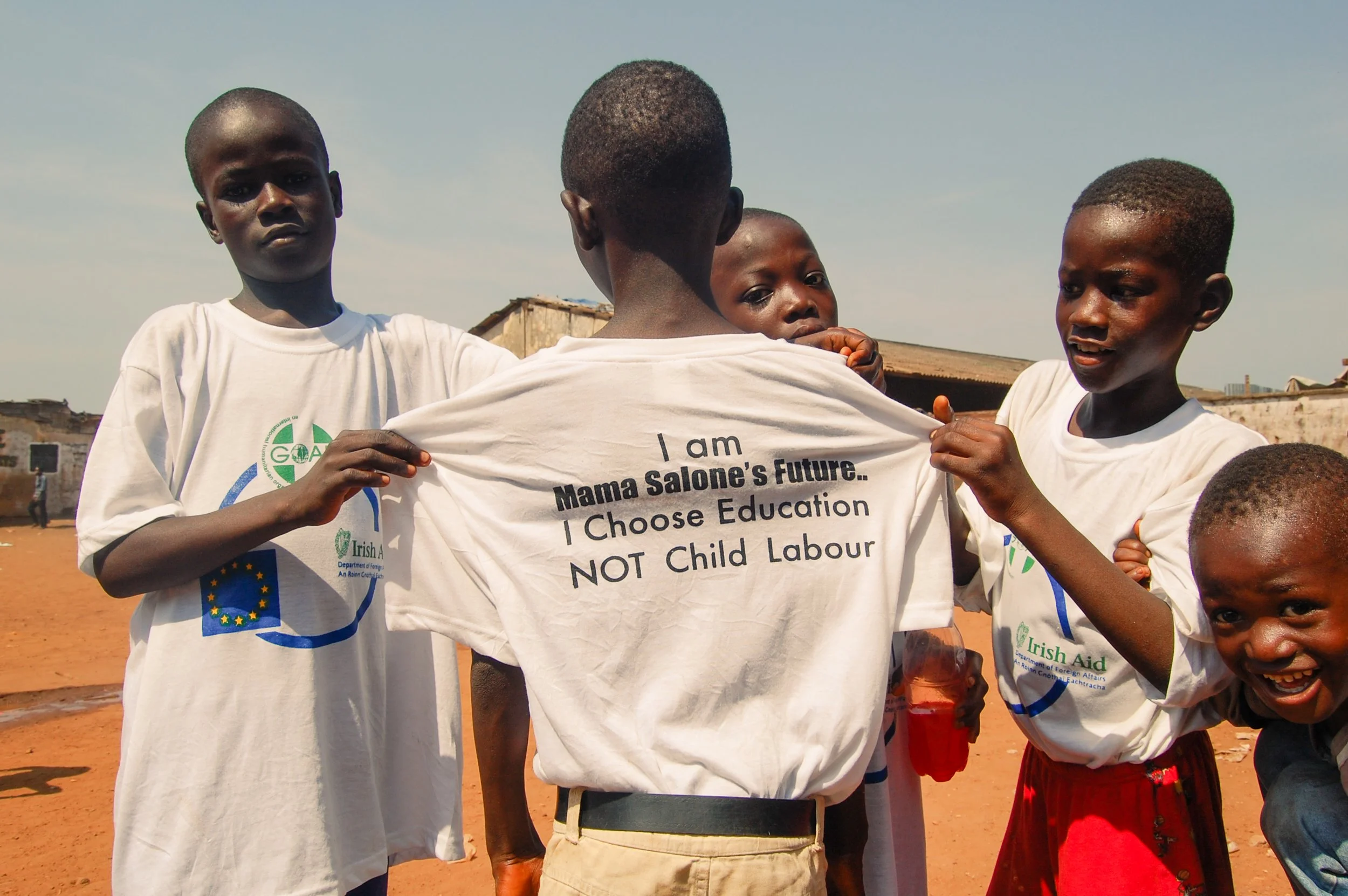 Verschillende kinderen die T-shirts dragen met teksten over kinderarbeid en onderwijs, buiten op een stoffige plek onder een heldere hemel.