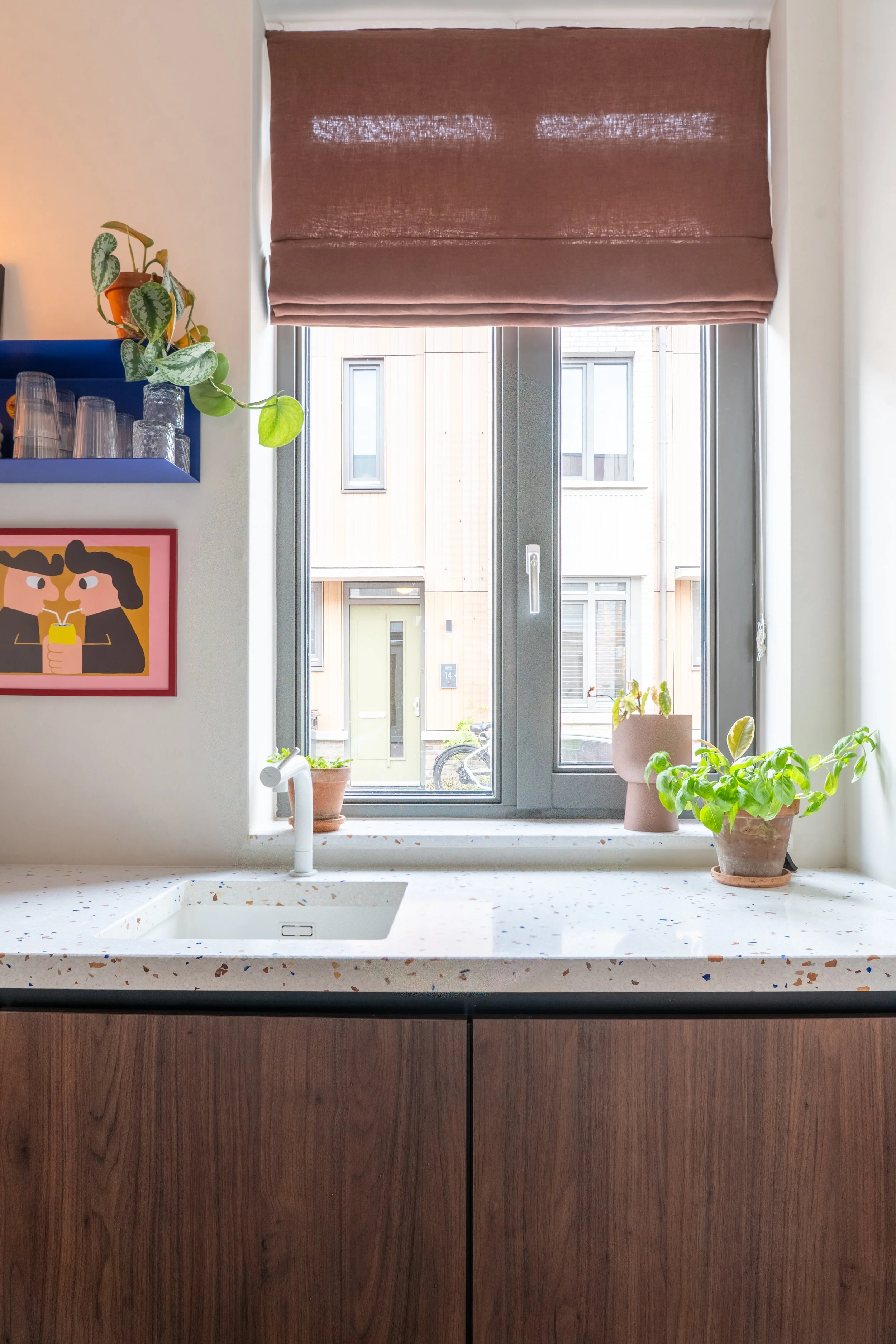 Keuken met een wit aanrecht en een wit wasbakje onder een raam met een rolgordijn. Op het aanrecht staan twee potten met planten en een muur met een kunstwerk. Buiten het raam zijn modern uitziende gebouwen te zien.