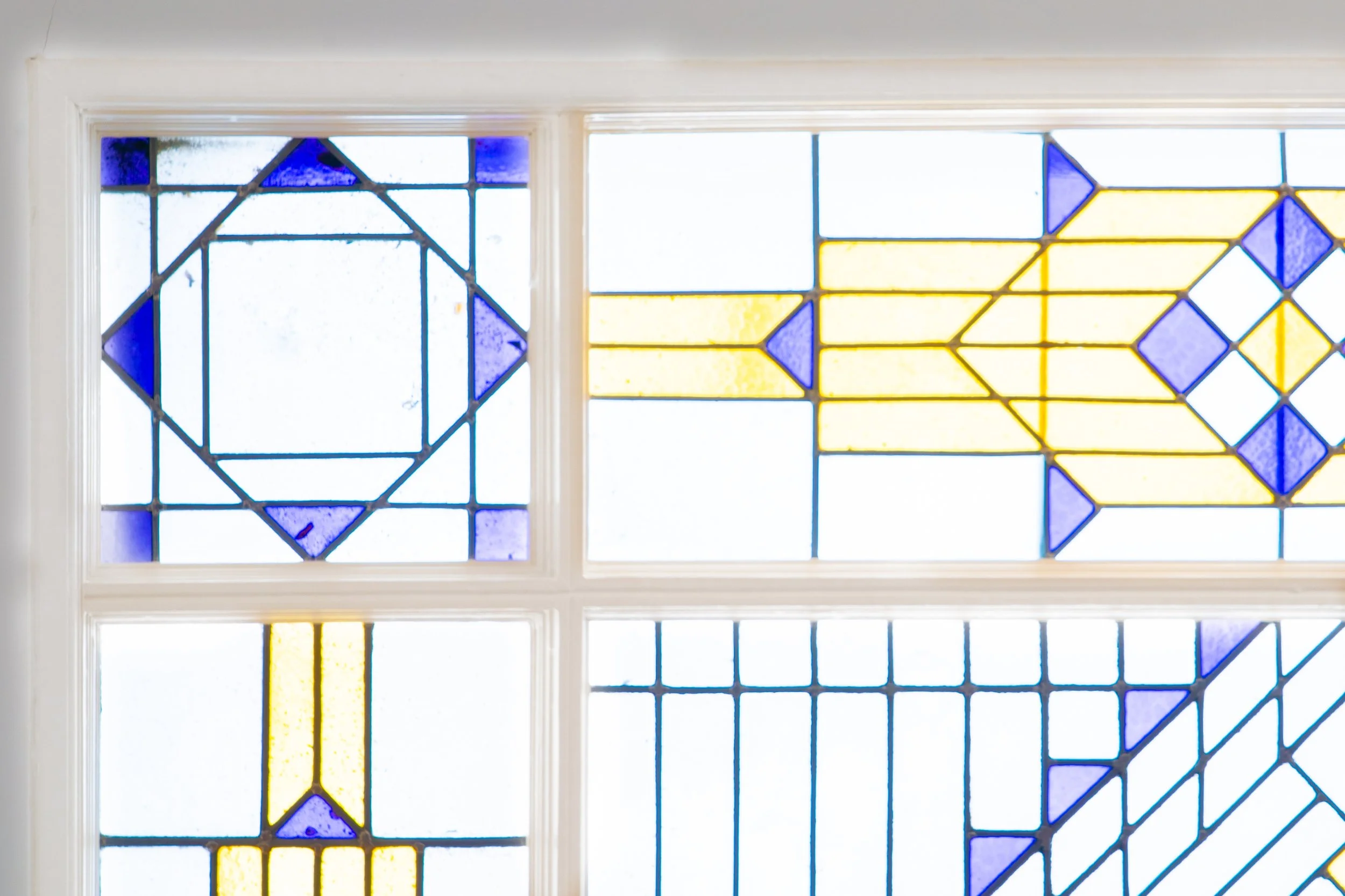 Kleurrode glas-in-lood ramen met geometrische patronen in blauw, geel en wit, ingelijst in wit kader.