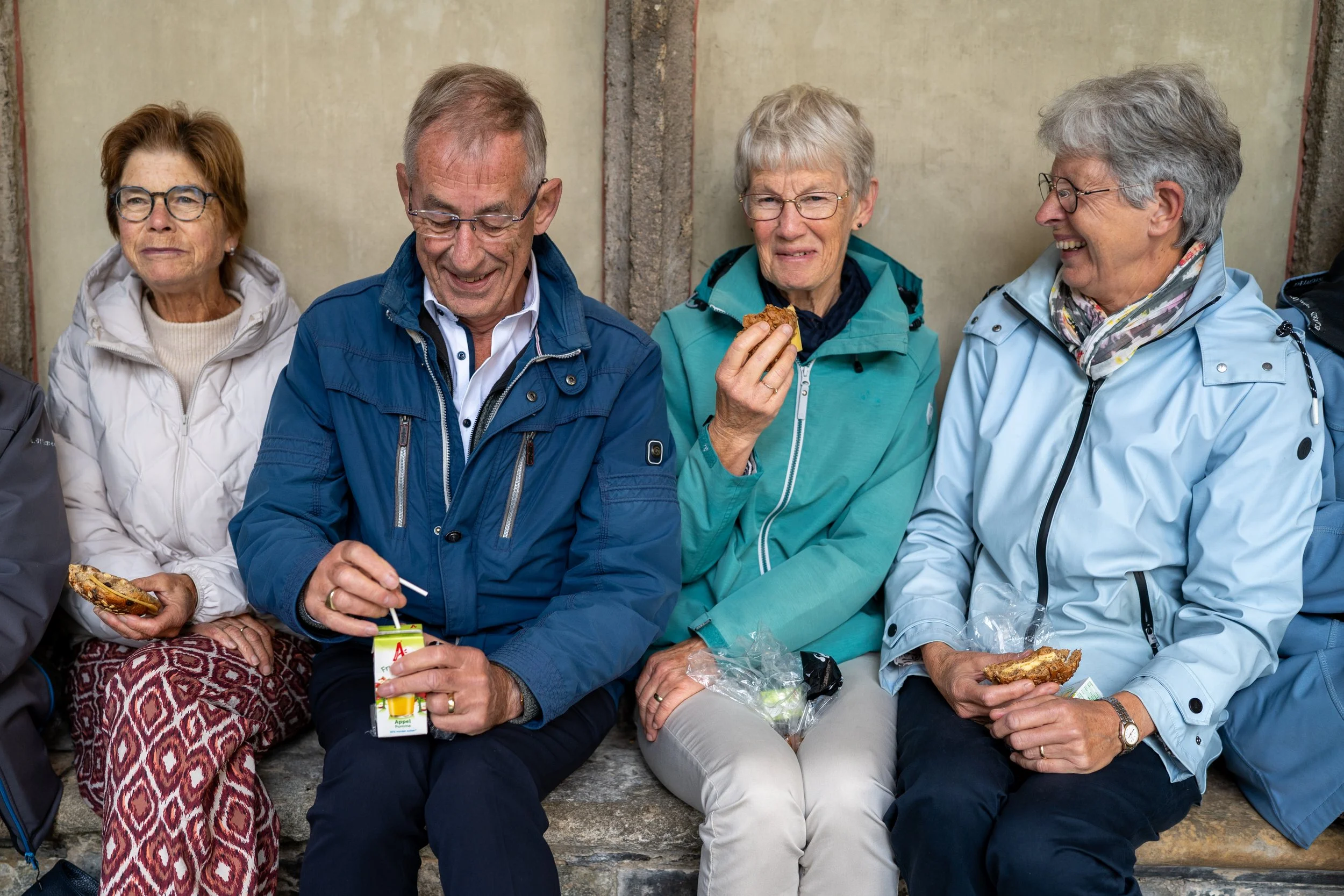 Vijf oudere mensen zitten samen buiten op een bankje, genieten van een gesprek en het eten, met een muur op de achtergrond.