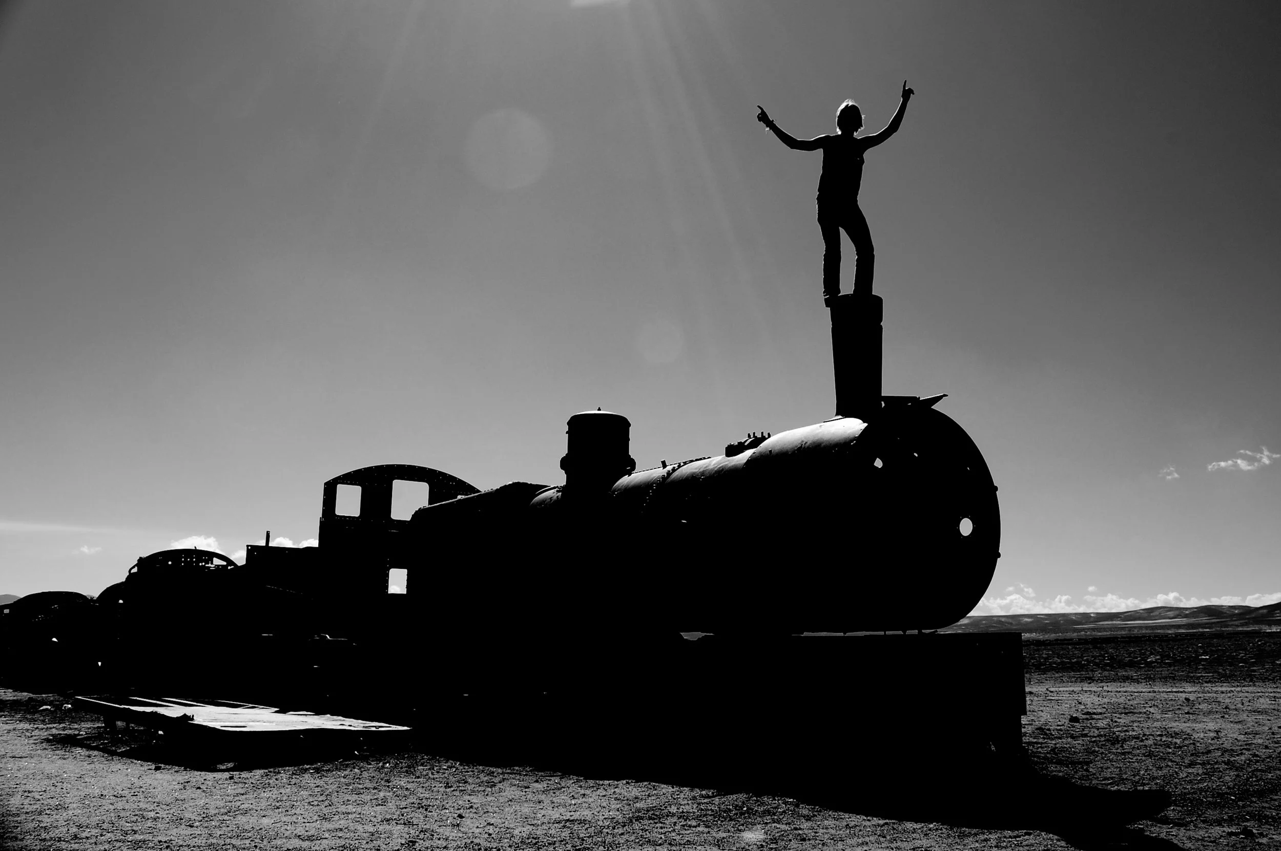 Silhouet van een persoon die op een oud, roestig vliegtuig staat, met de armen in de lucht, tegen een heldere hemel met de zon die schijnt.