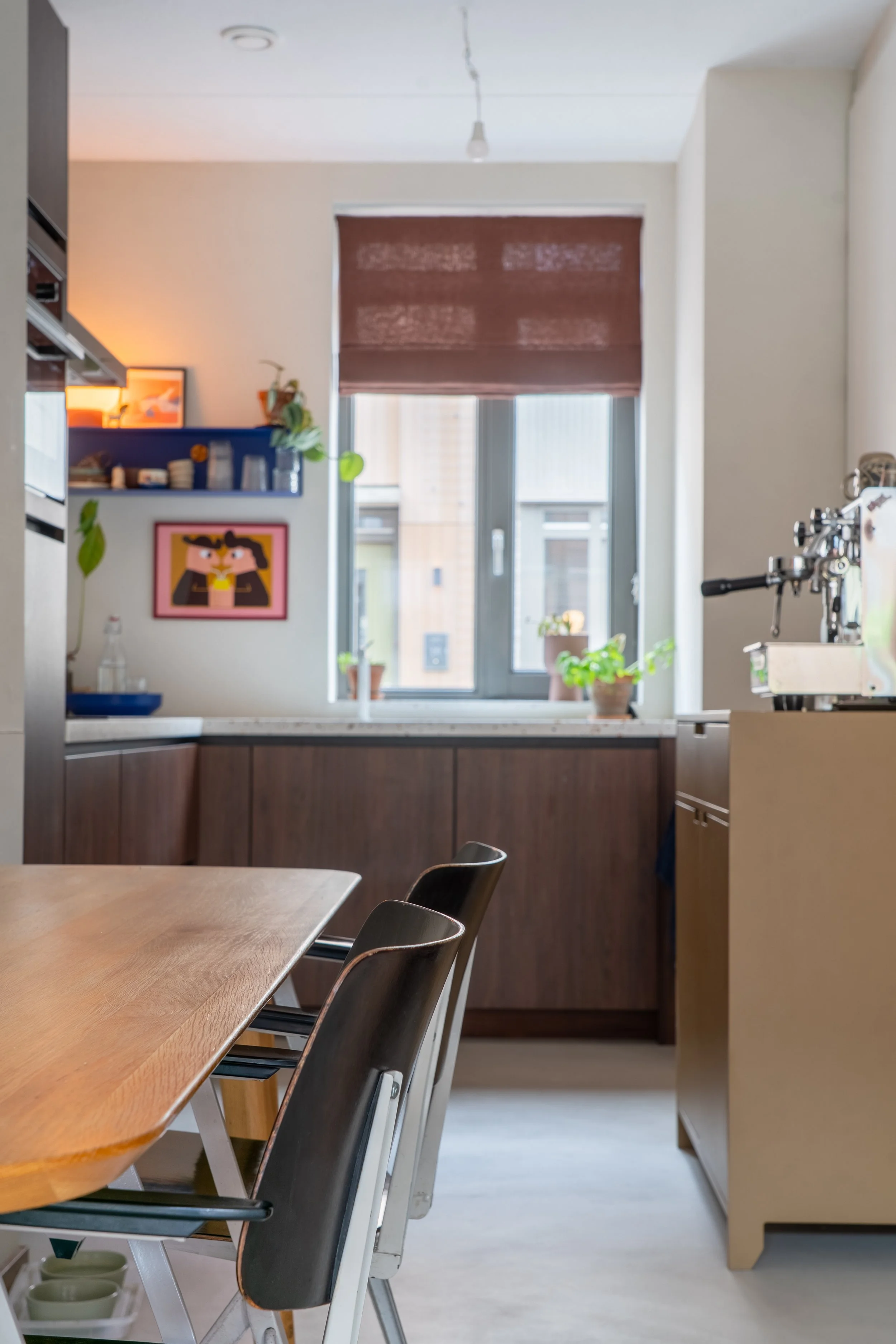 Een keuken met een houten eettafel en zwarte stoelen. In de achtergrond een raam met een rolgordijn en enkele planten op het raamkozijn, er is ook een waterkoker en andere keukenapparatuur te zien.