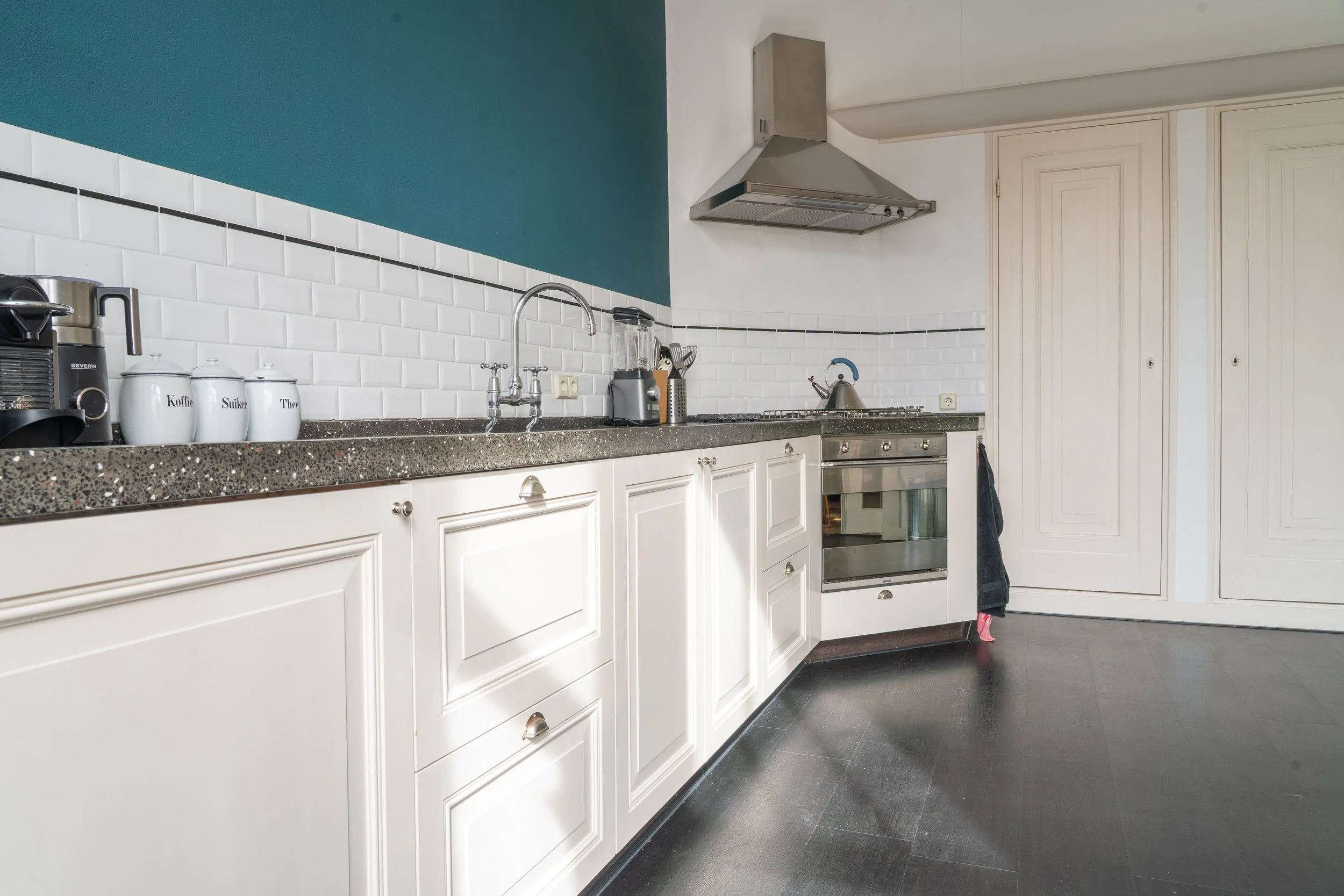 Keuken met witte kasten, een zwart steengoed werkblad, en een witte betegelde muur met zwarte lijsten. Er hangt een oven en een gasfornuis met een stoomkoker en een waterkoker. Aan de muur hangen elektrische stopcontacten en een blauwe kraan boven de