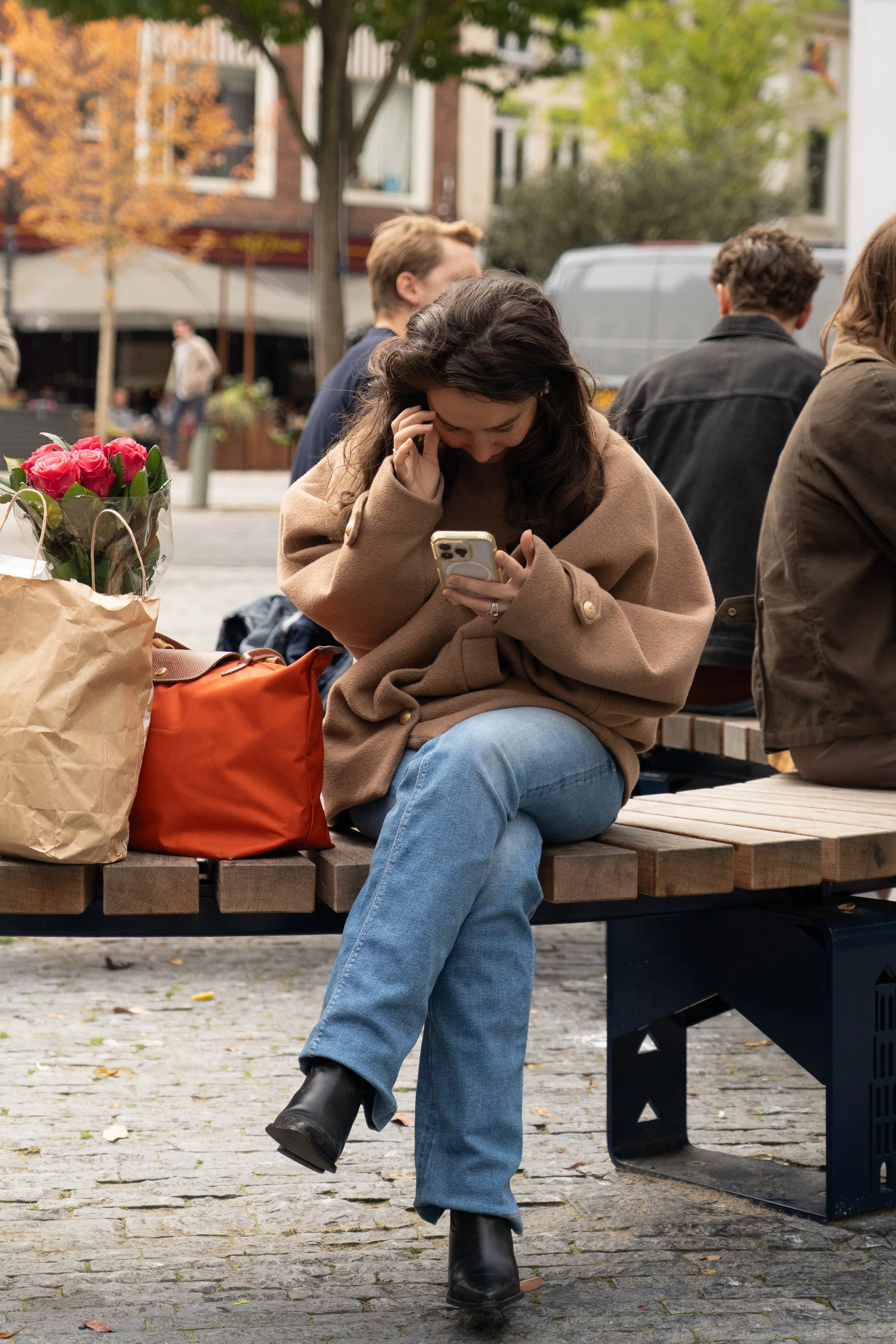 Een vrouw zit op een bank in een park, kijkt naar haar telefoon, met een bos bloemen en een oranje tas naast haar. Ze draagt een beige jas, blauwe spijkerbroek en zwarte laarzen. Om haar heen zitten andere mensen op bankjes, en in de achtergrond zijn