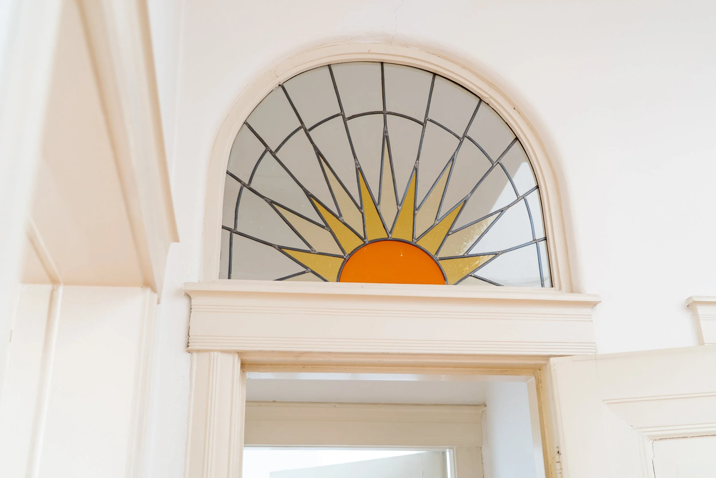 Een halfcirkelvormig raam in een witte muur met een zonnestraal design in gekleurd glas, boven een witte omlijsting.