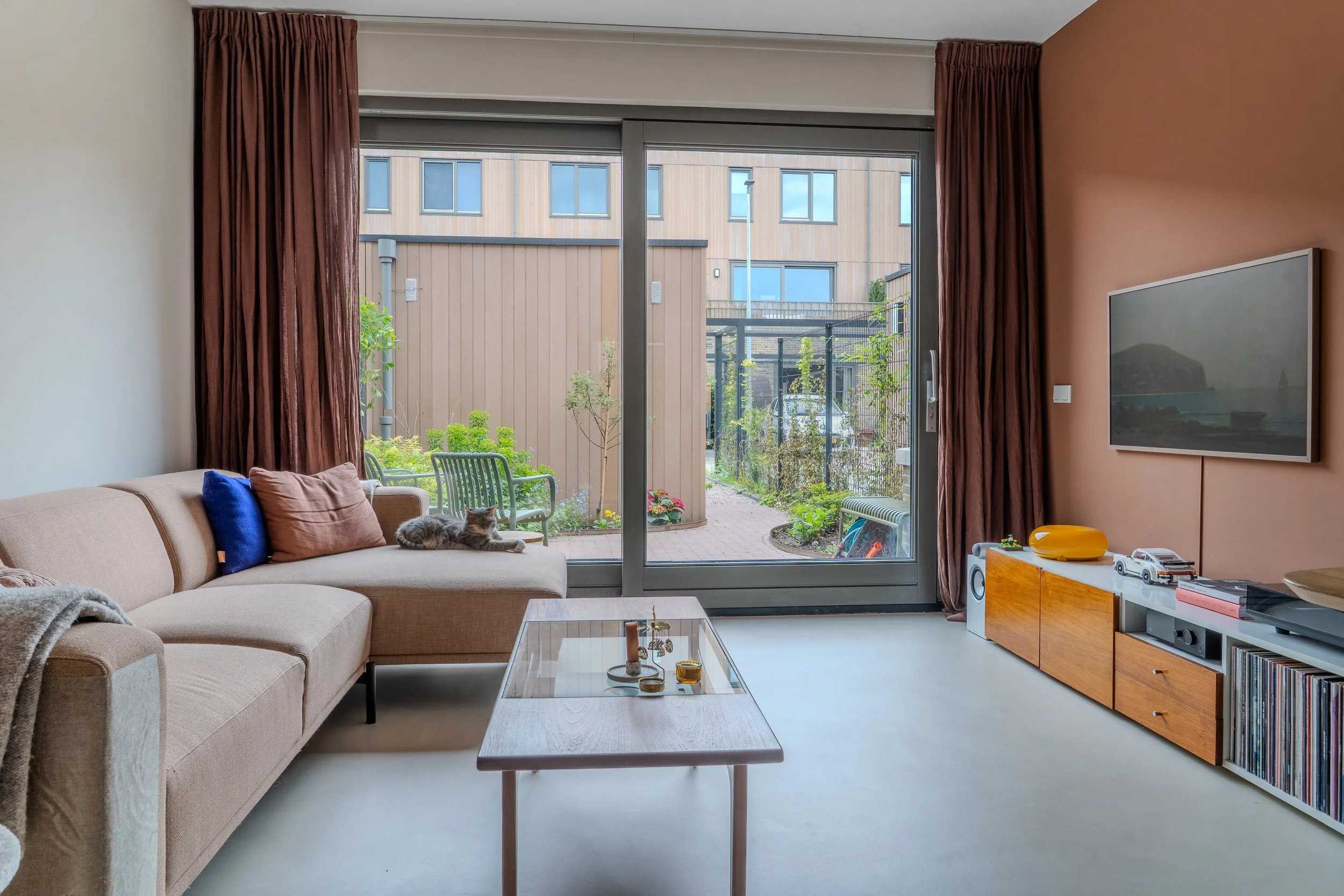 Woonkamer met grote ramen, bank met kussens, katten op de bank, salontafel met decoratie, tv aan de muur, houten kast met boeken, speelgoed op de kast.