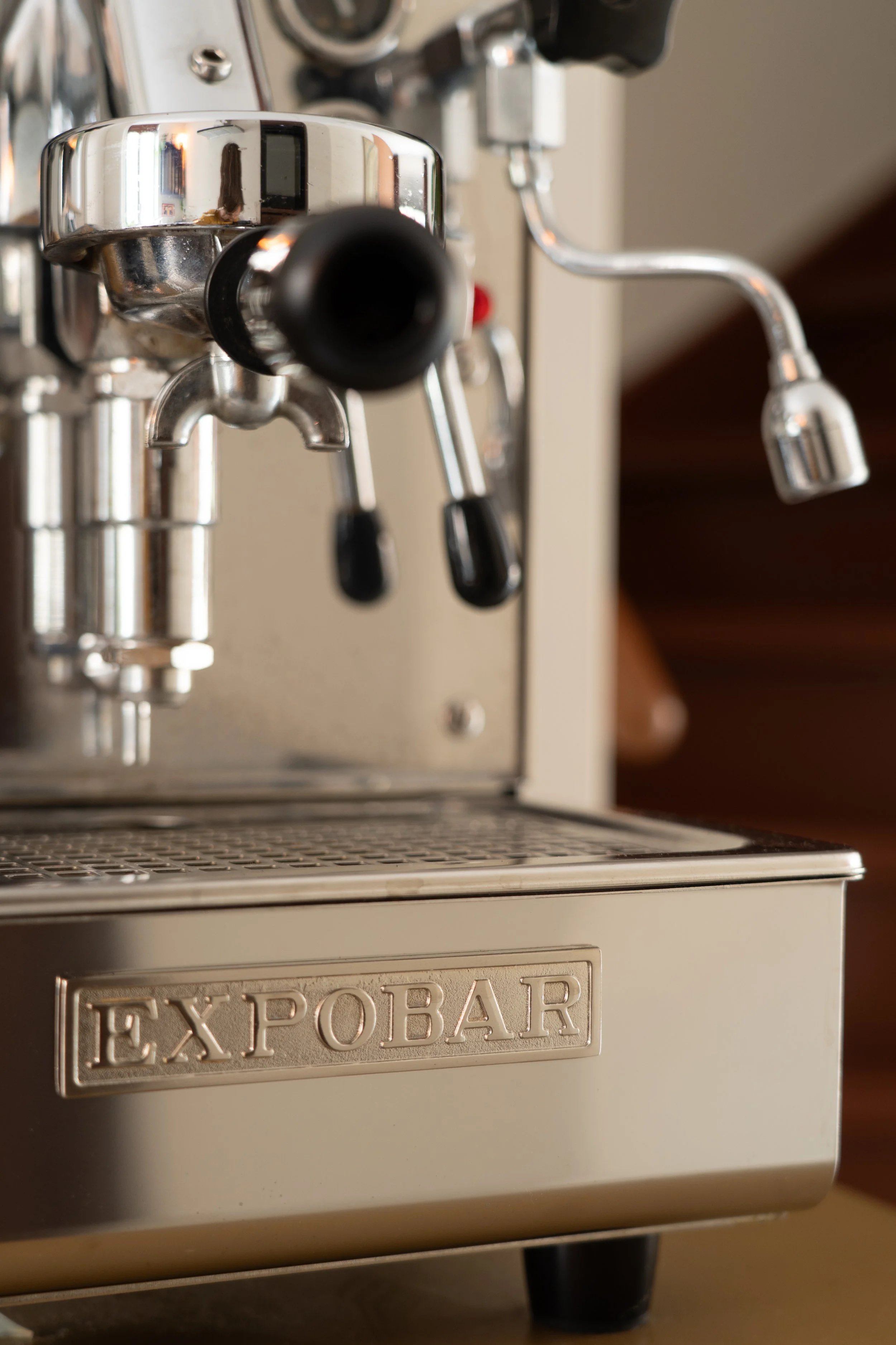 Close-up van een espressoapparaat met de merknaam 'EXPOBAR' op de voorkant, metalen onderdelen en een zwart uitloopstuk.
