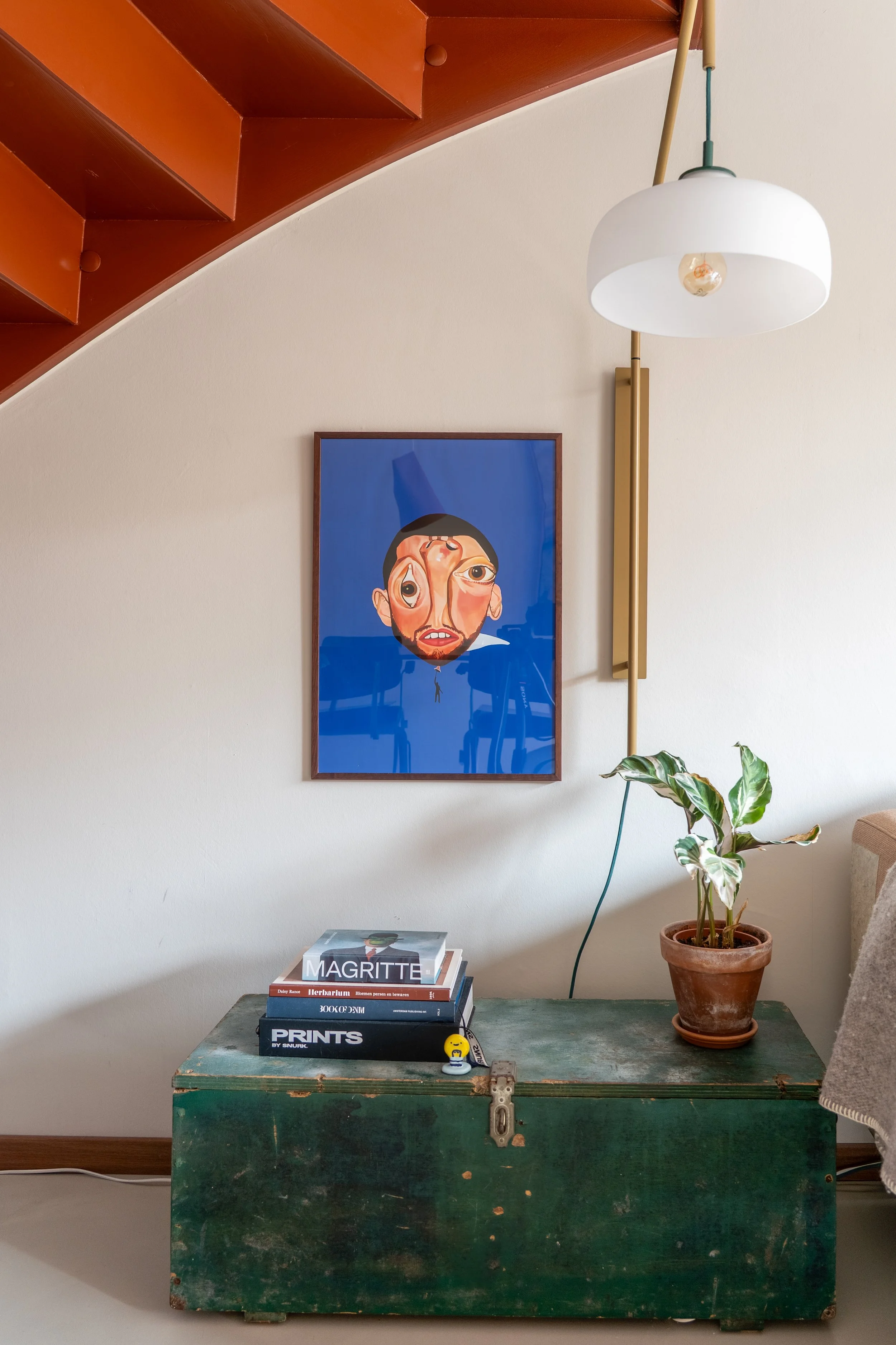 Een vintage groene houten kist met diverse boeken en een kleine knuffel op de voorkant, naast een potted plant op een tafel, tegen een witte muur met een kunstwerk en een wandlamp.