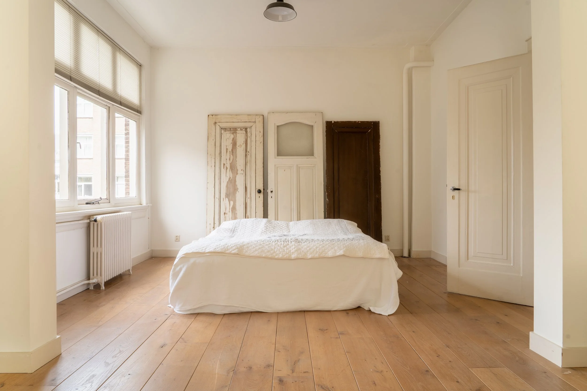 Een slaapkamer met houten vloeren en witte muren. Een bed met een witte deken staat onder twee oude, weerwinkende deurpanelen die tegen de muur zijn geplaatst. Aan de linkerkant een raam met luiken en een radiator. Boven het bed hangt een plafondlamp