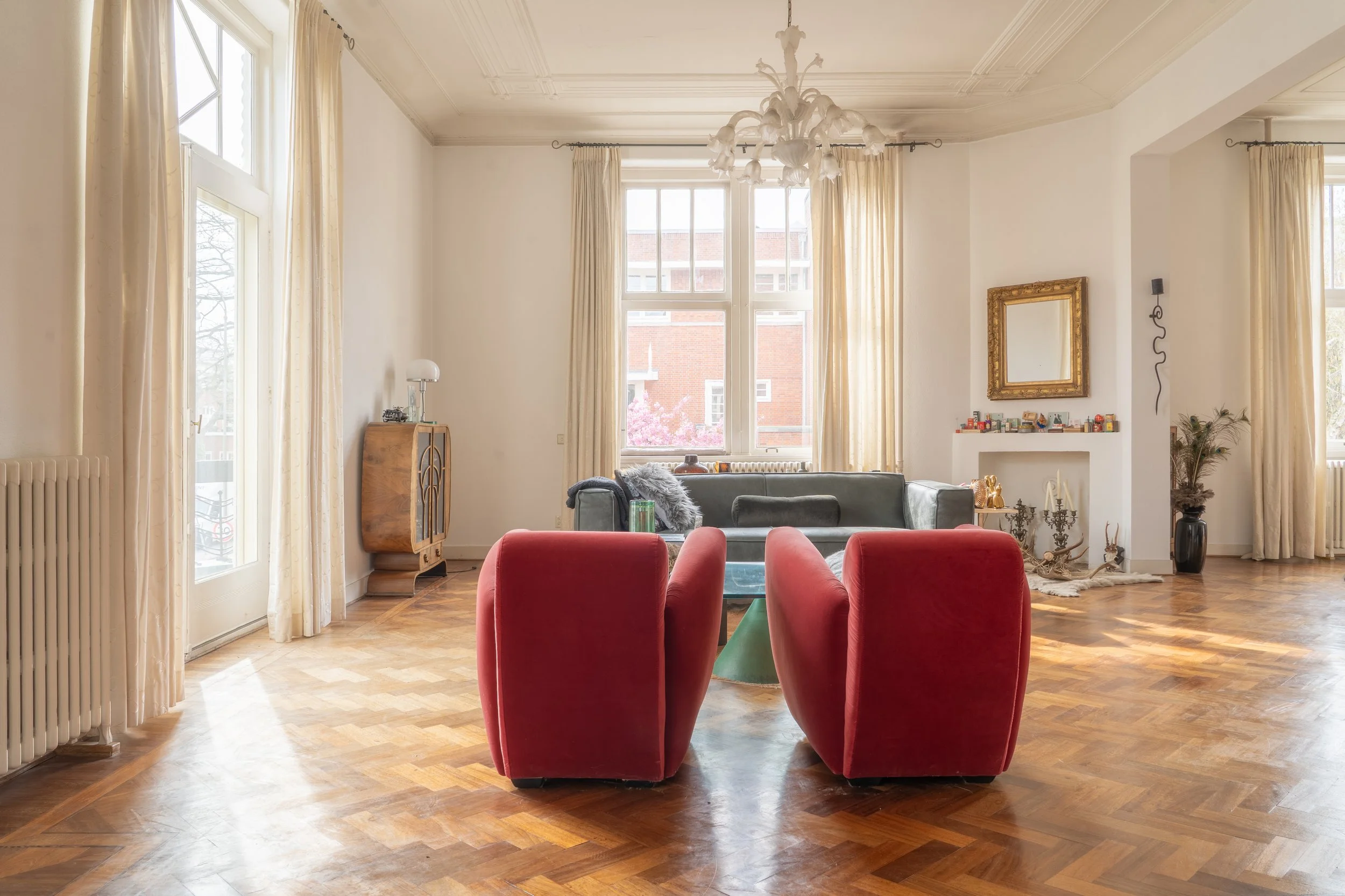 Een woonkamer met grote ramen, beige gordijnen, een chandelier, een blauwe bank, rode stoelen, een spiegel met een gouden lijst, en verschillende decoraties zoals planten en kandelaars.